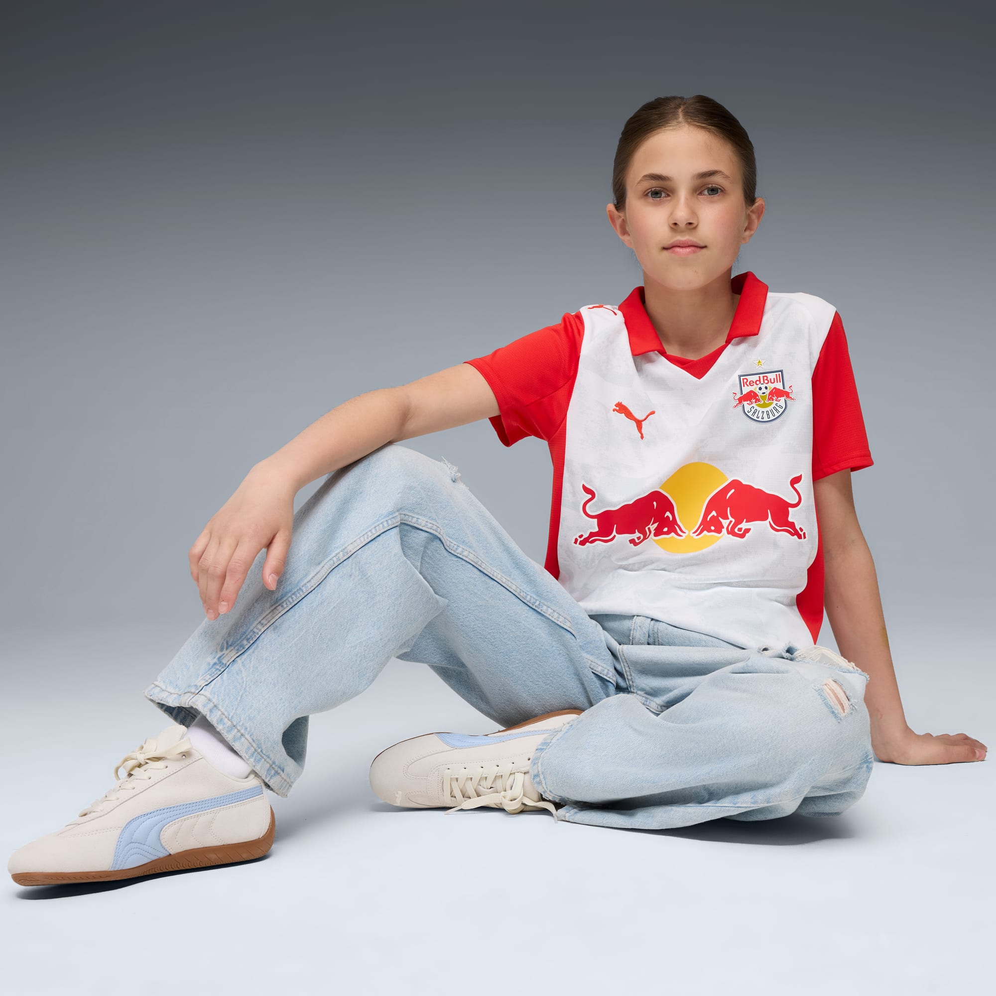 Ein Model sitzt vor grauem Hintergrund, mit Jeans, Turnschuhen und rotem Red Bull Shirt.