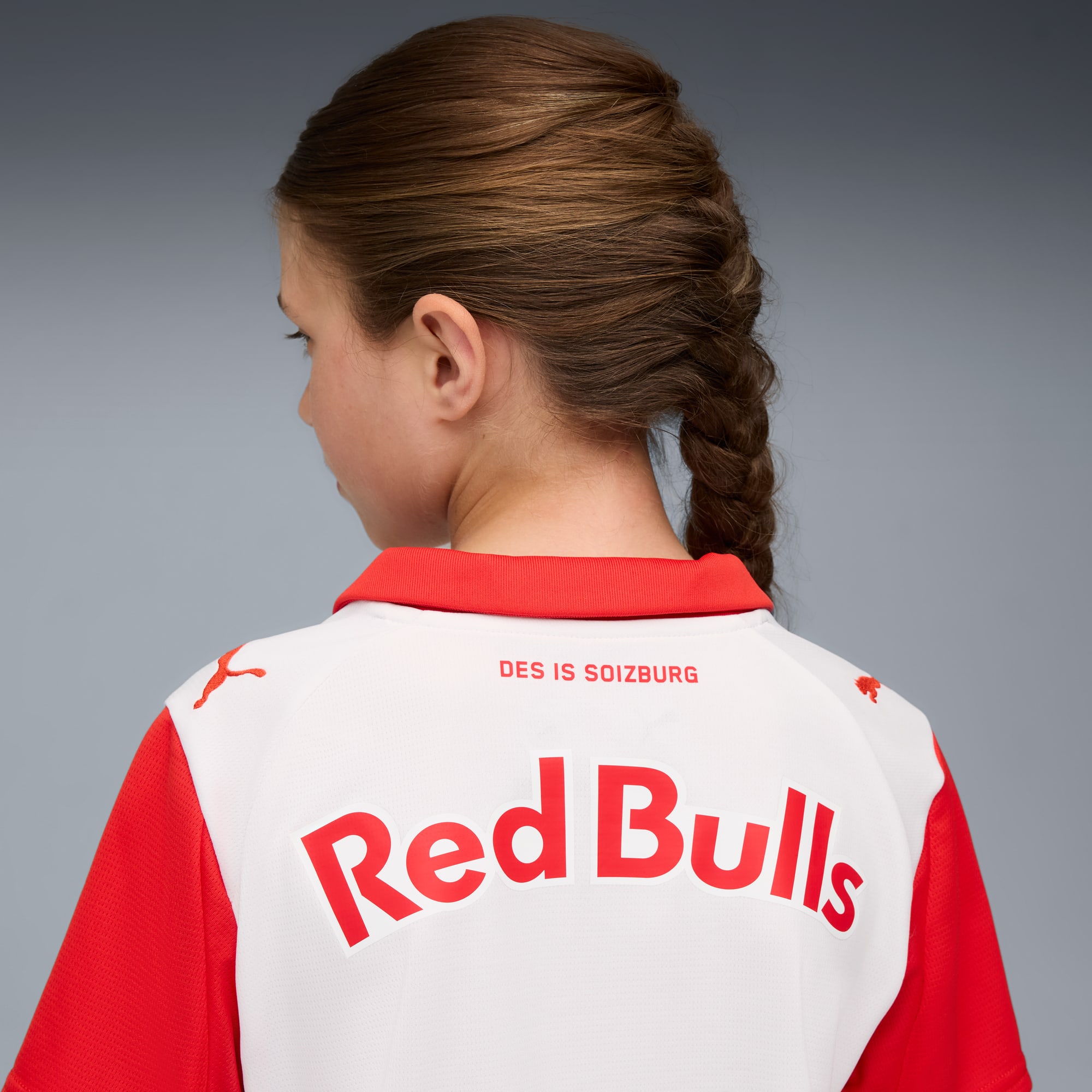Vor grauem Grund trägt ein Mädchen ein rot-weißes 'Red Bulls'-Shirt, von hinten.