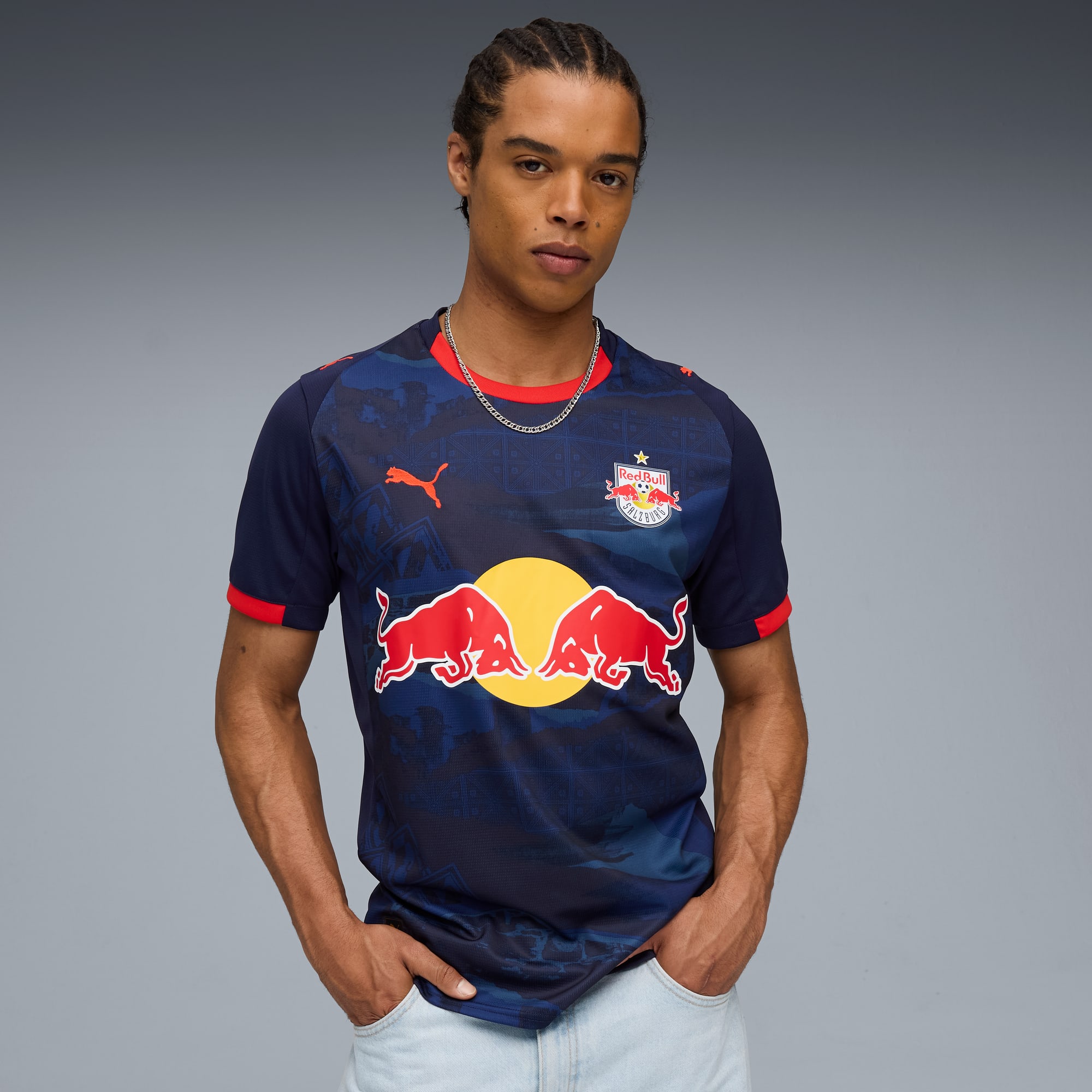 Een model poseert tegen een grijze achtergrond, gekleed in een blauw Red Bull Salzburg T-shirt.