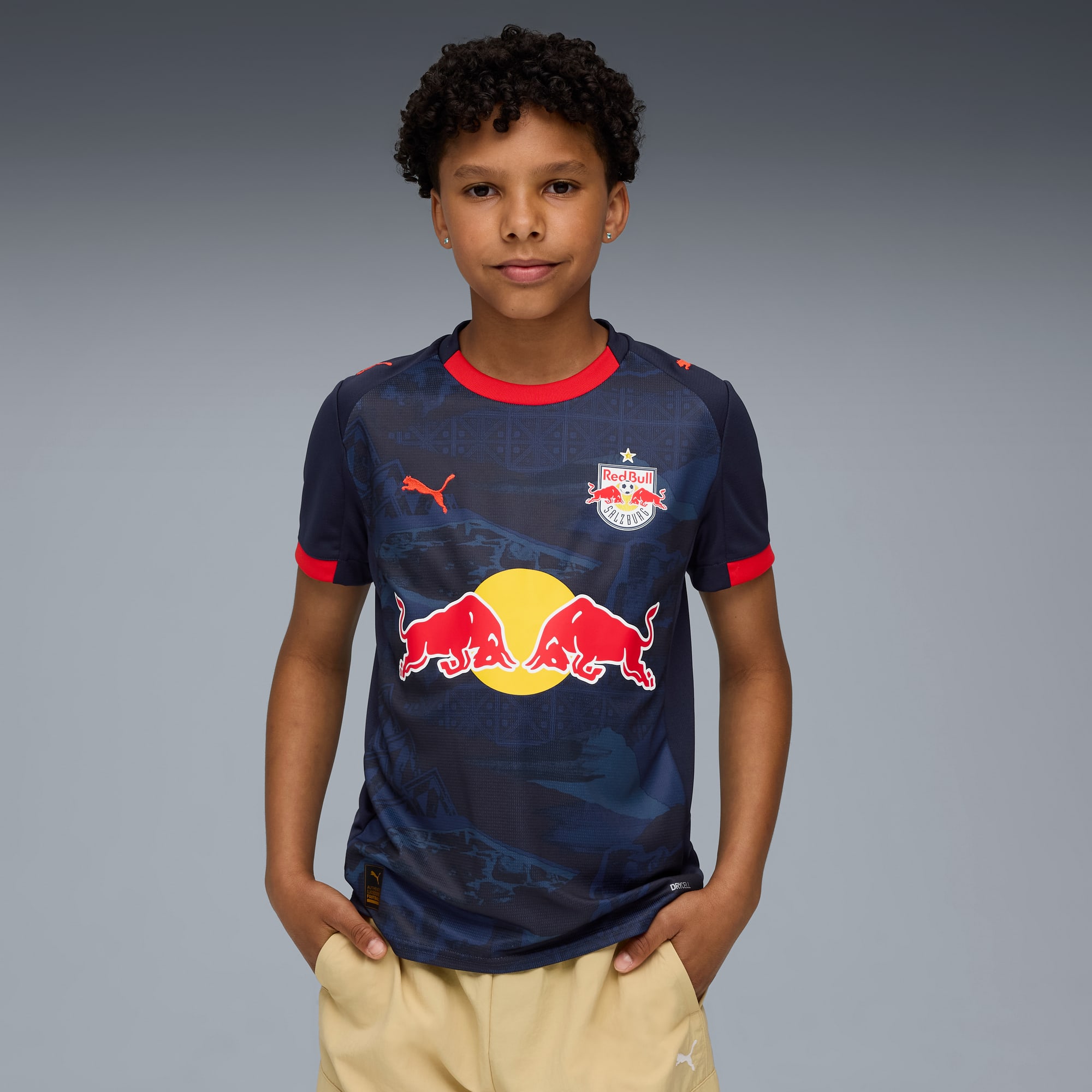 Ein Kind posiert vor grauem Hintergrund in einem marineblauen Fussballtrikot mit roten Details.