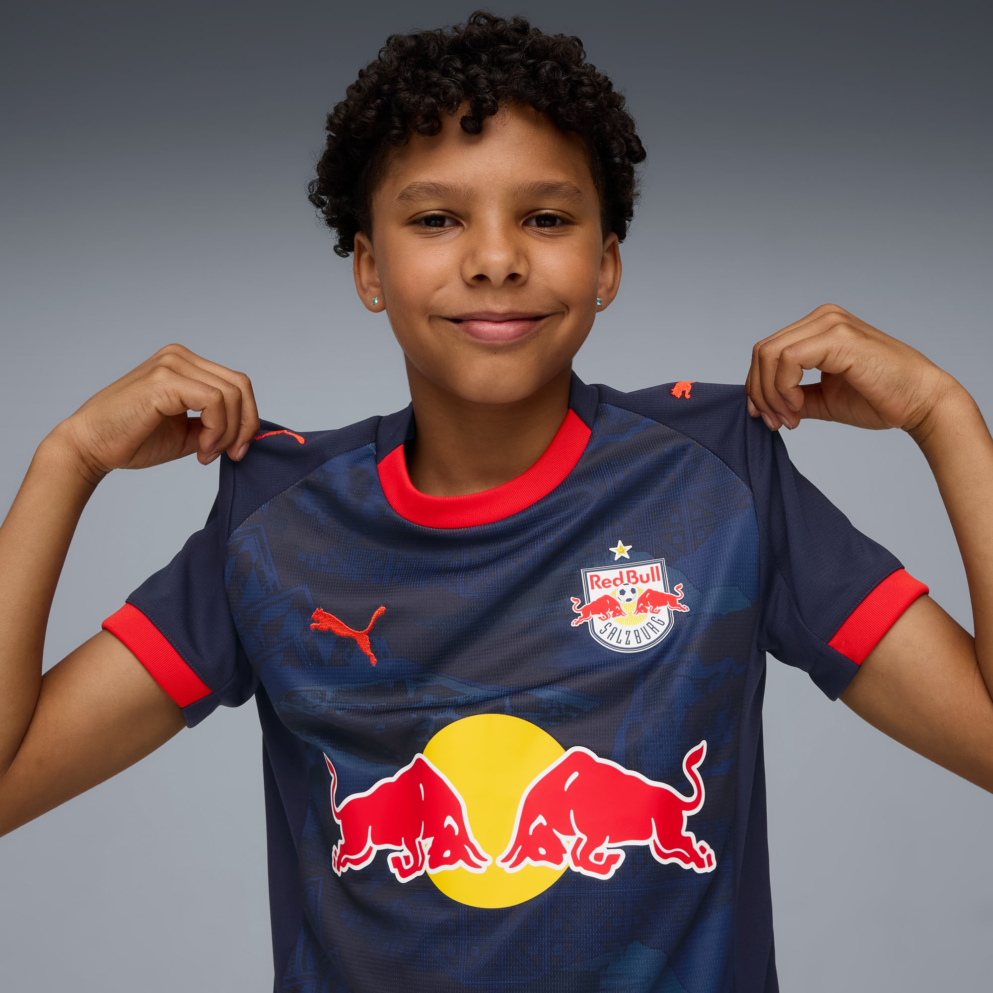 Vor grauem Hintergrund lächelt ein Model und präsentiert ein kurzärmeliges Sportshirt mit Logo.