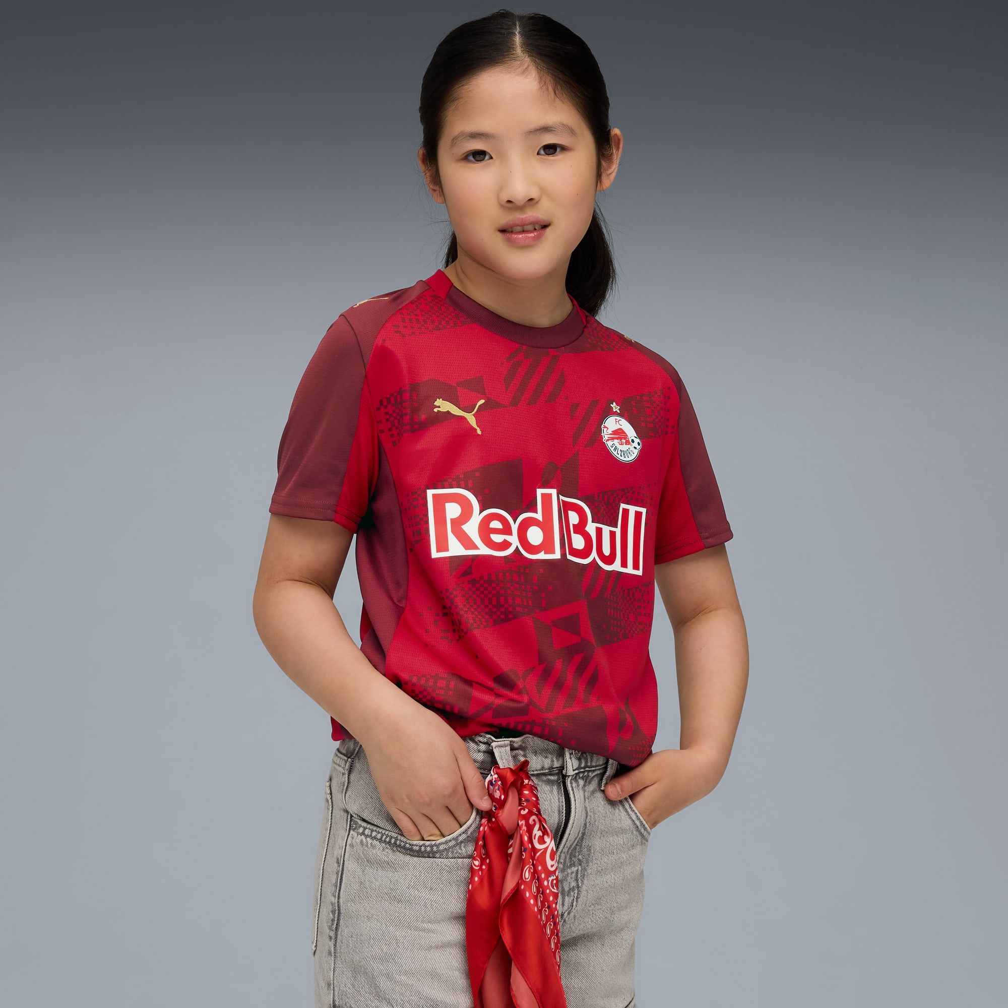 Vor grauem Hintergrund posiert ein Model in rotem Red Bull T-Shirt und grauen Shorts.