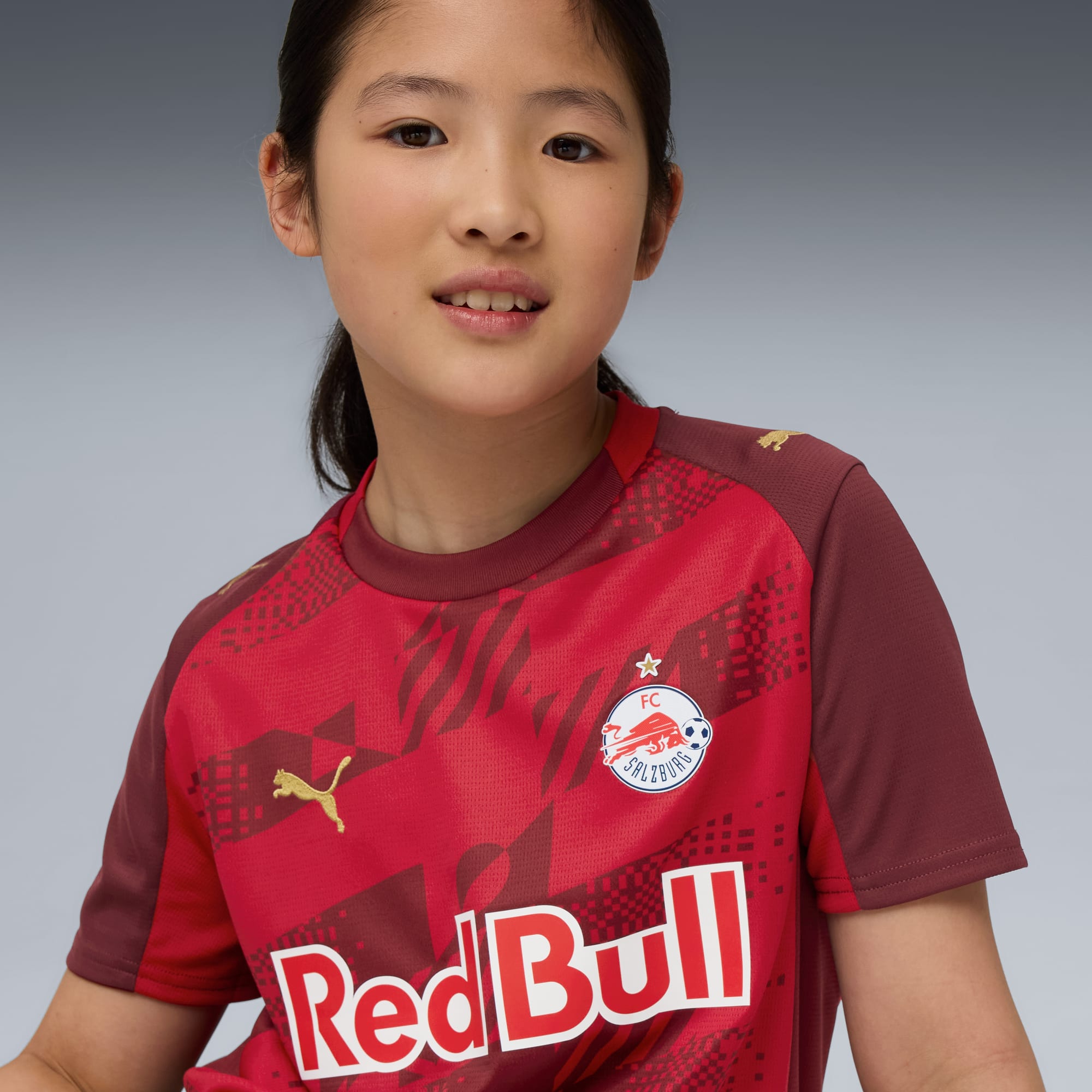 Vor grauem Hintergrund posiert ein Model in einem roten FC Salzburg T-Shirt mit 'Red Bull'-Text.