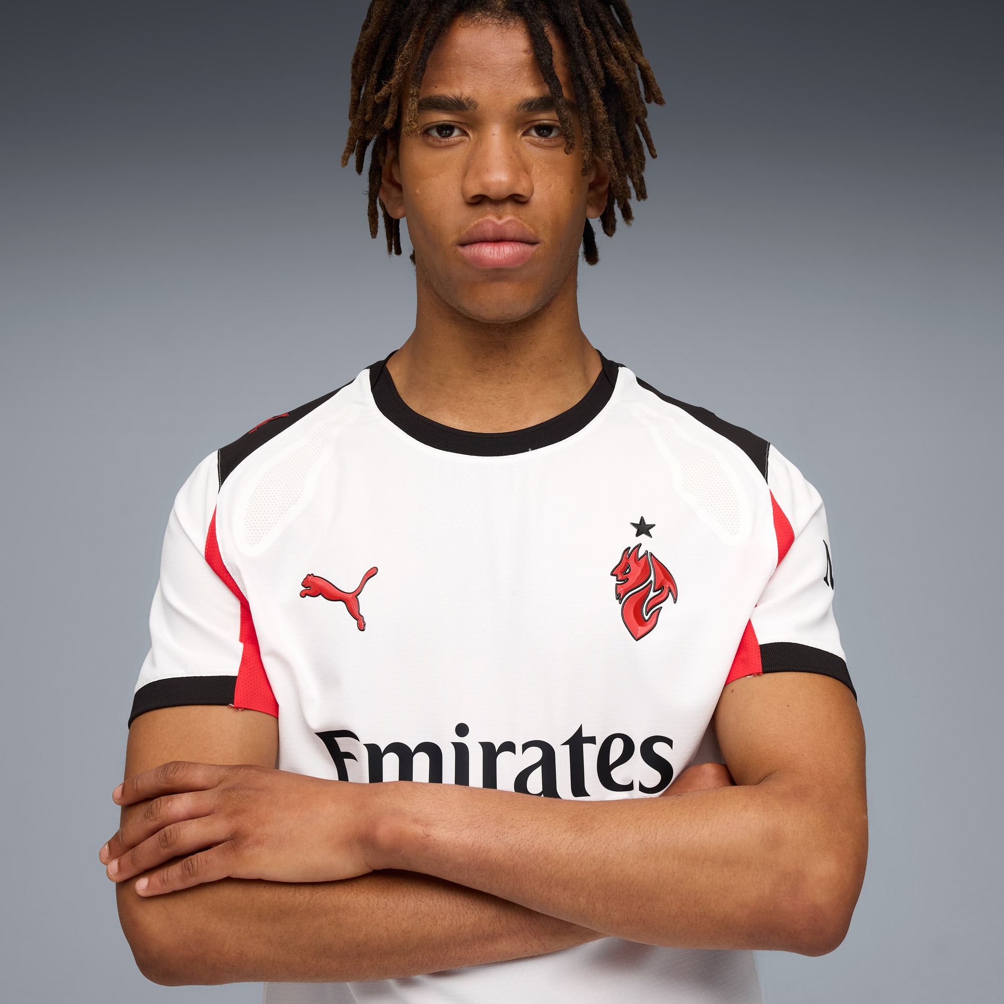 Un modello con dreadlocks posa su sfondo grigio indossando una maglia sportiva bianca.