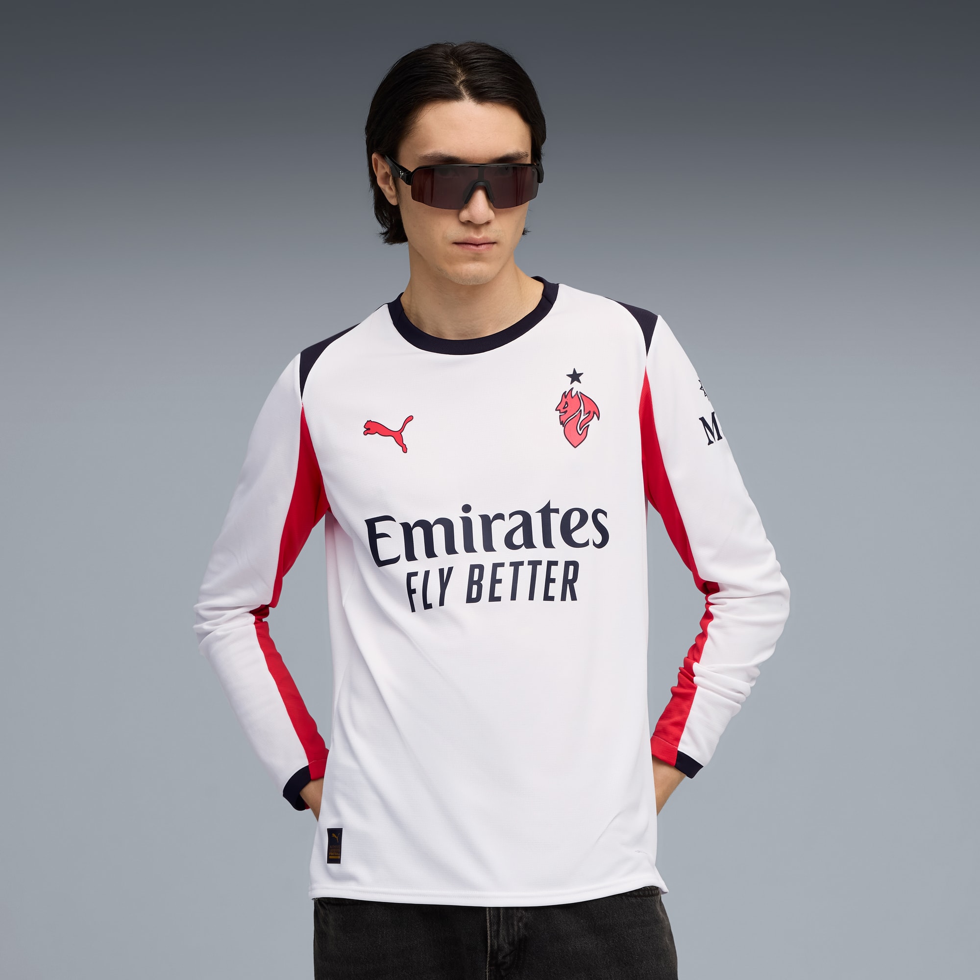 Sur fond gris, un mannequin porte des lunettes de soleil et un t-shirt blanc à manches longues avec des détails rouges et marine.