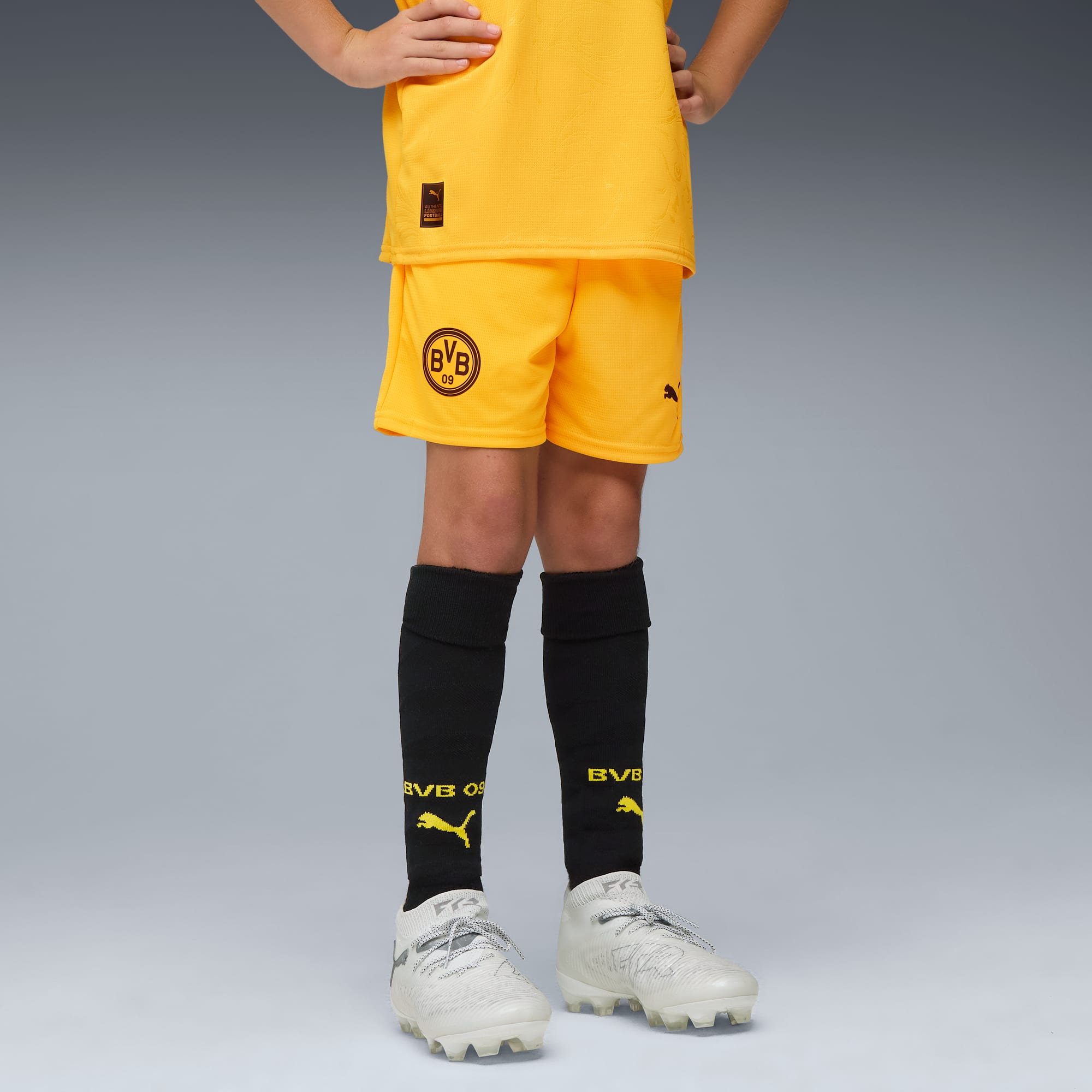 Model na szarym tle ma na sobie żółty strój, czarne skarpety i białe buty piłkarskie.