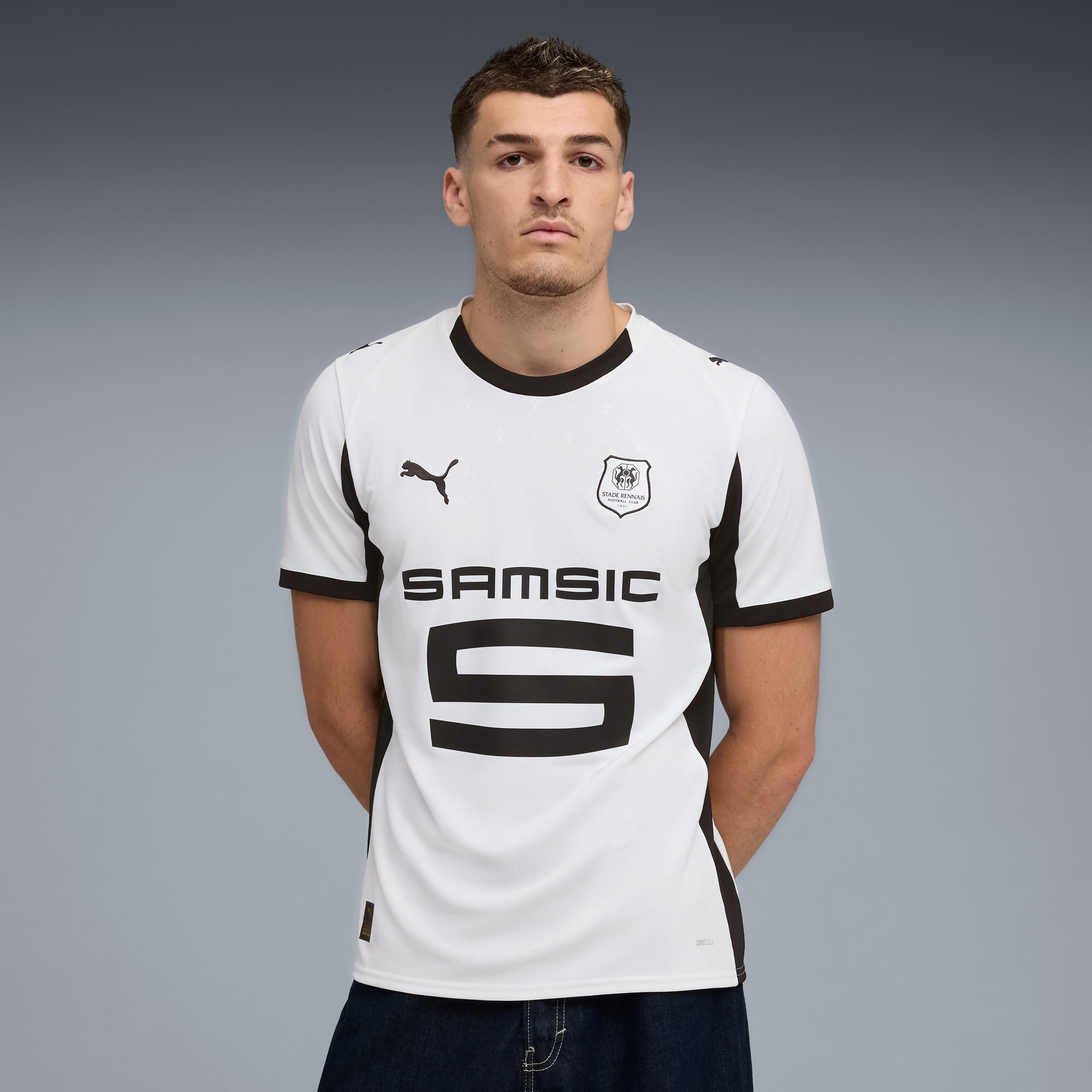 Een model poseert tegen een grijze achtergrond, gekleed in een wit shirt met zwarte accenten en donkere jeans.
