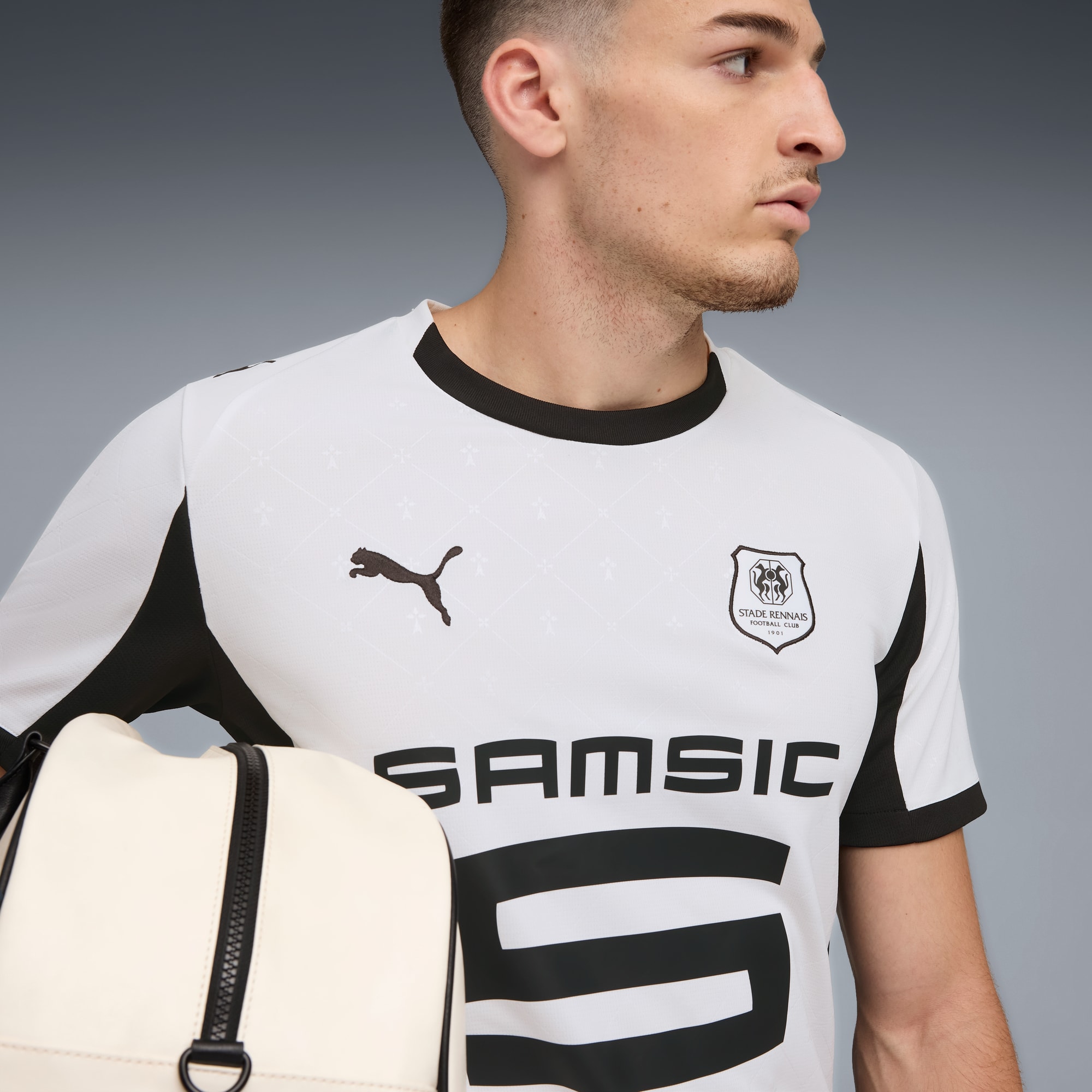 Tegen een grijze achtergrond draagt een model een wit voetbalshirt en een sporttas.