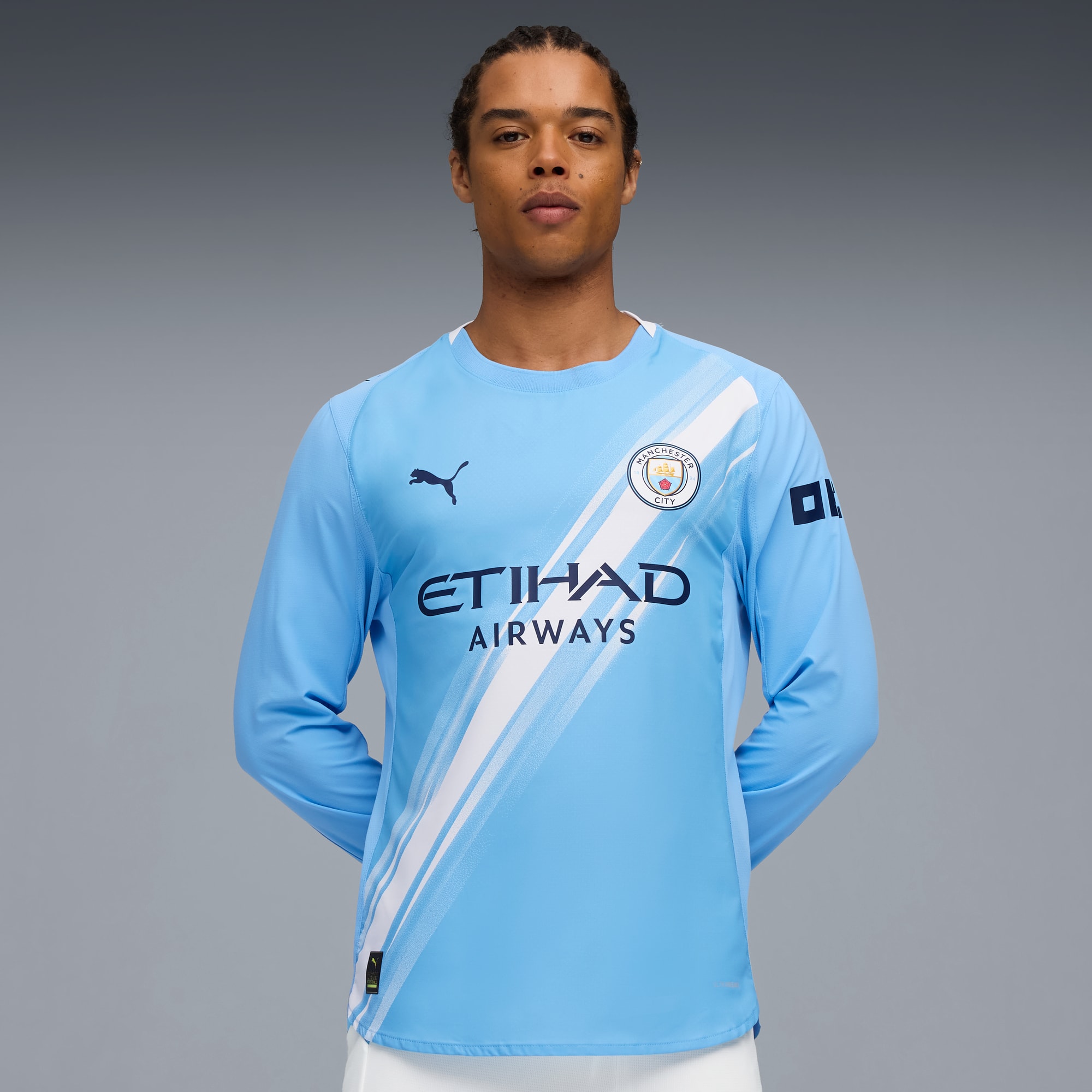 Against grey, a model wears a light blue, long-sleeved sports shirt, posing with arms behind.