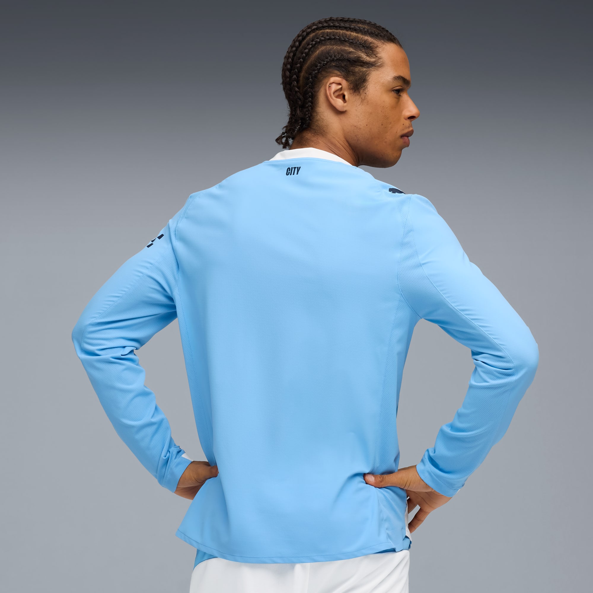 Against grey, a model with braided hair poses in a light blue, long-sleeved shirt.
