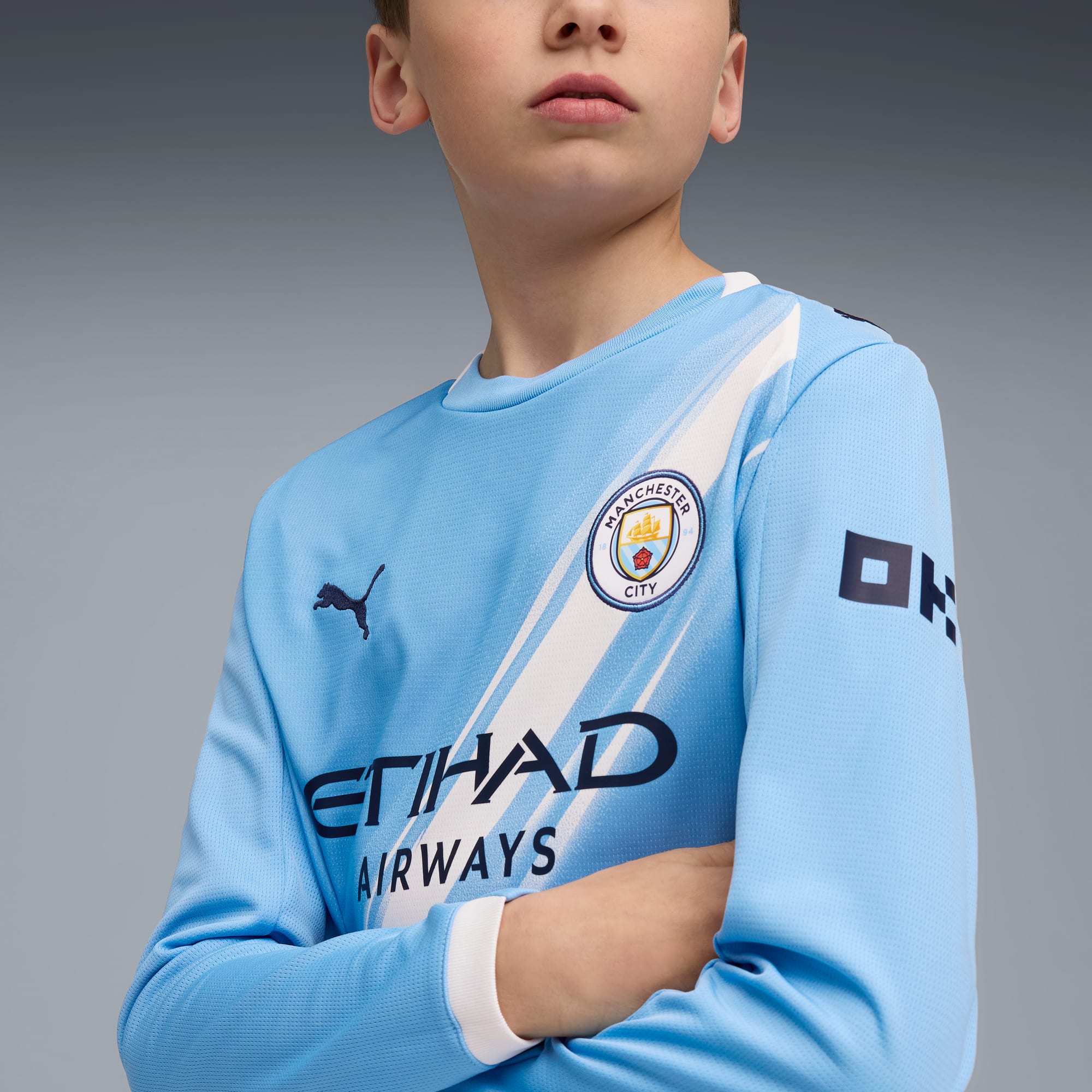Against grey, a model poses in a light blue, long-sleeved Manchester City football shirt.
