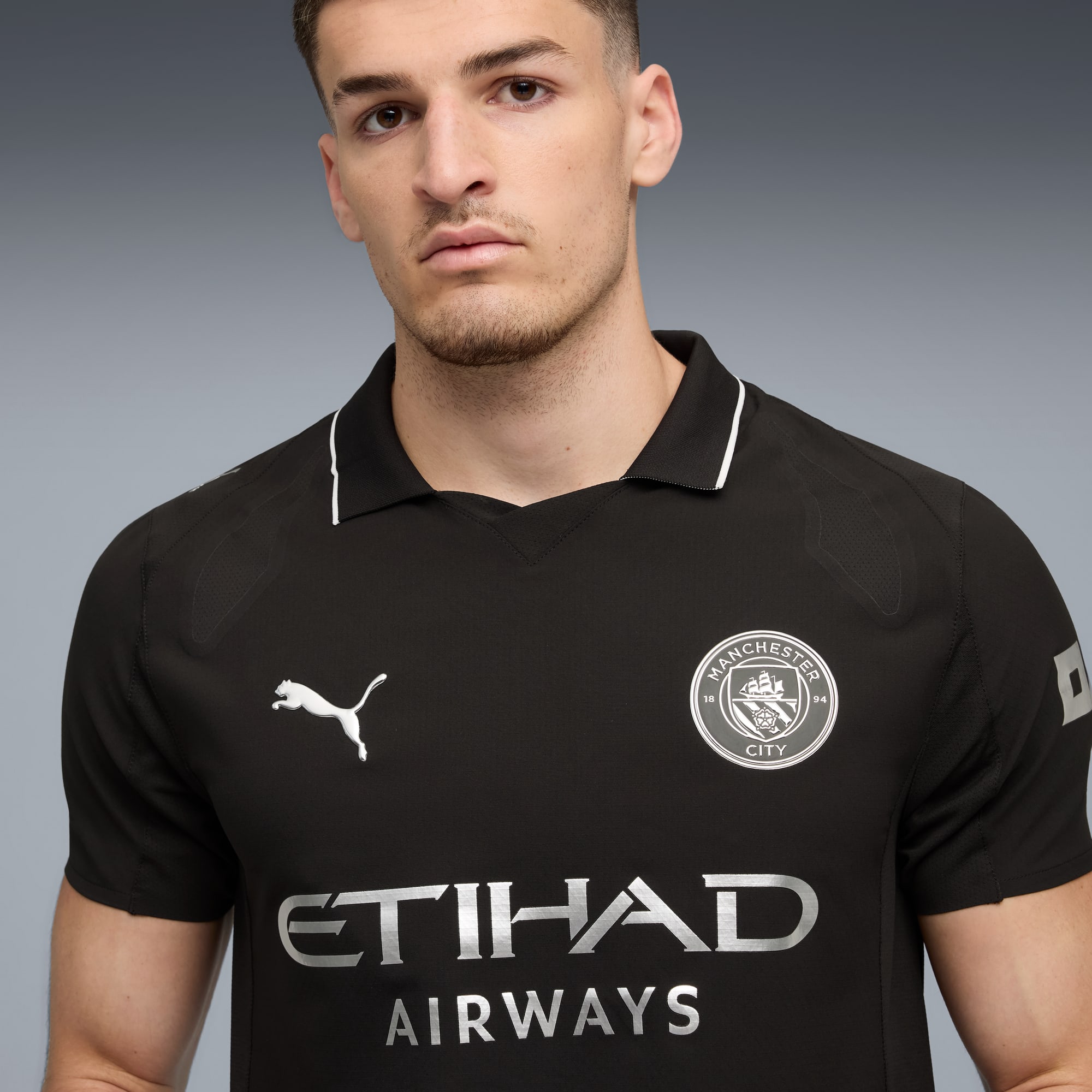 Un modello posa su sfondo grigio indossando una maglia nera del Manchester City.