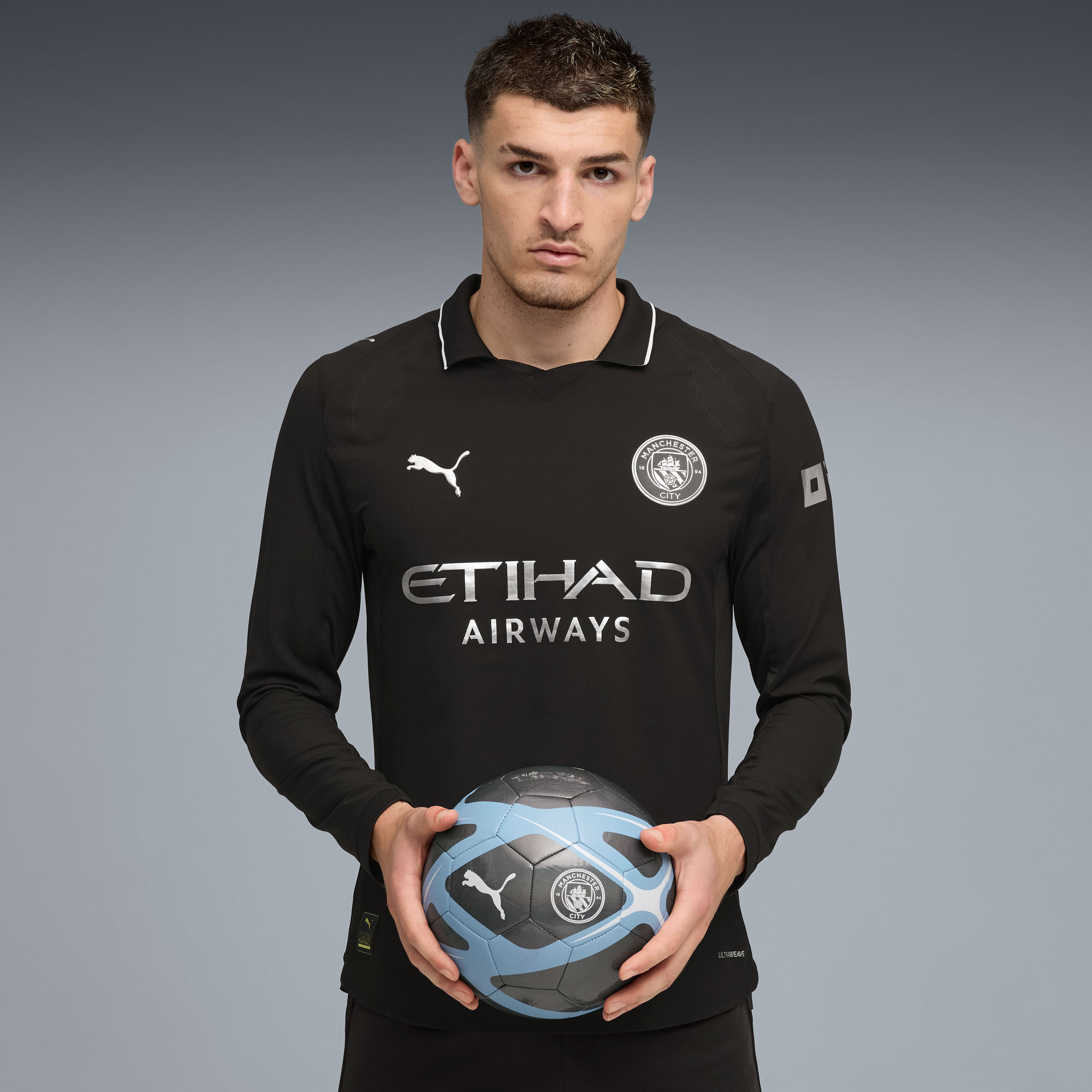 Sur fond gris, un modèle tient un ballon de foot et porte un haut noir à manches longues.