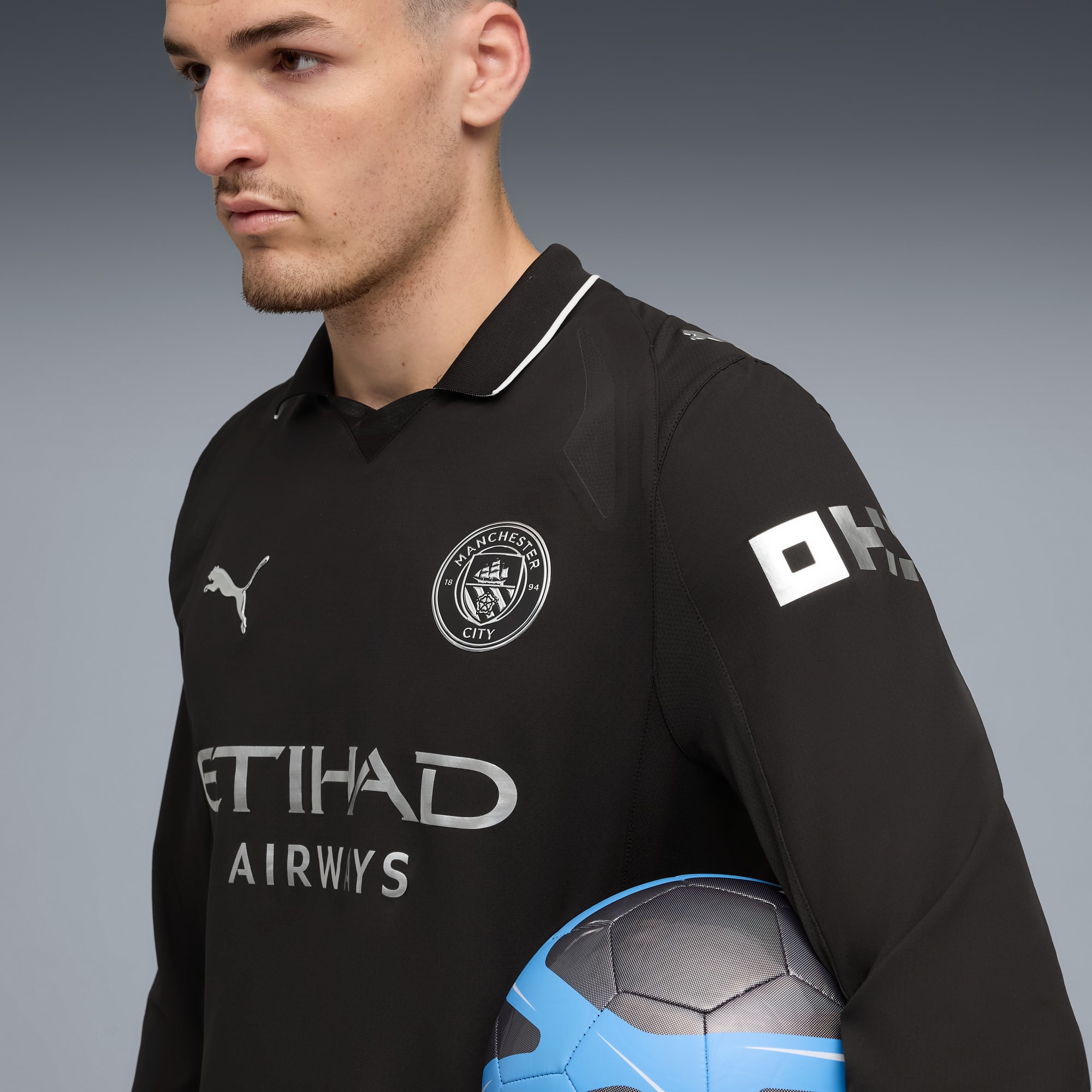 Un mannequin tient un ballon sur fond gris, portant un maillot noir à manches longues de Manchester City.
