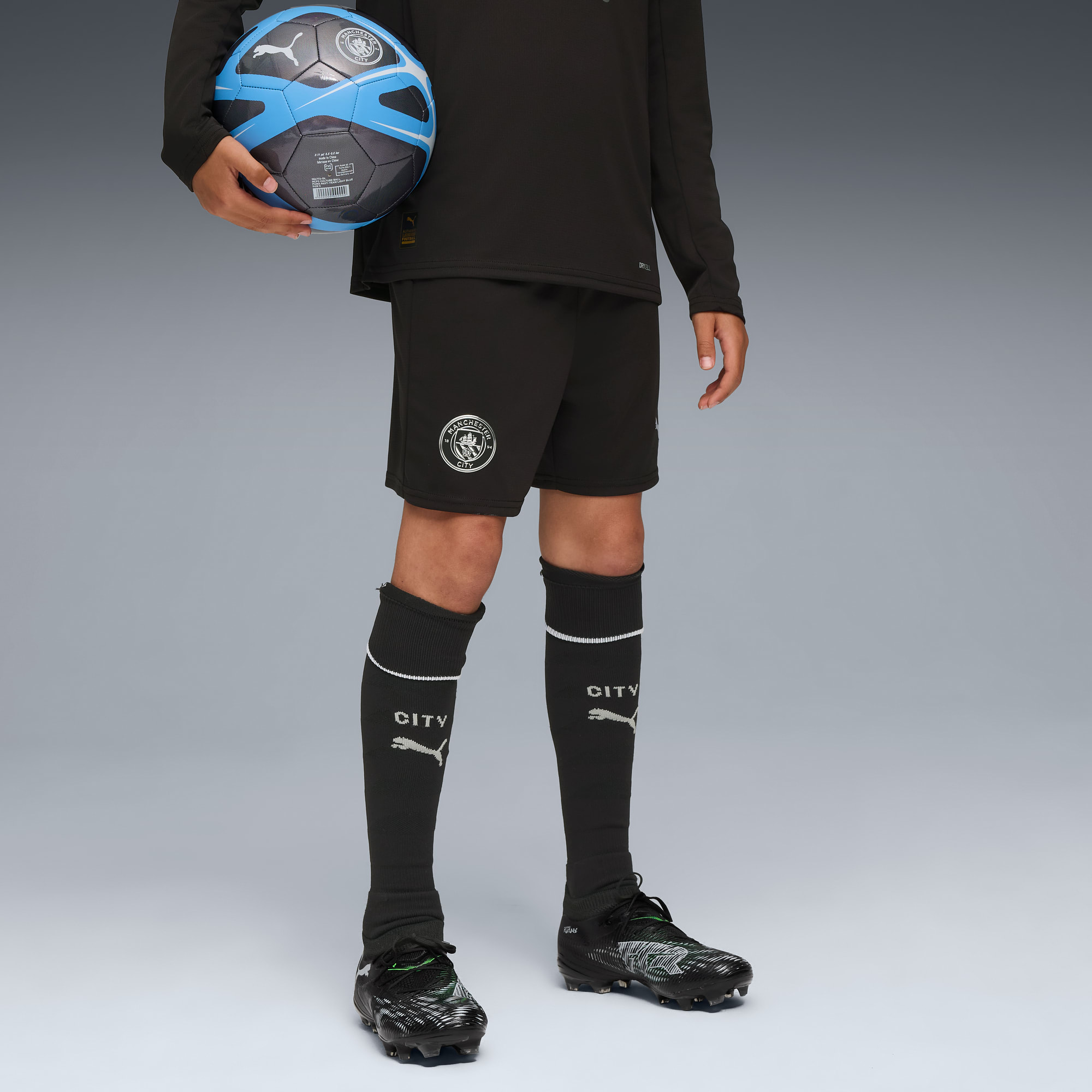 Sur fond gris, un modèle tient un ballon, vêtu d'un haut, short, chaussettes et chaussures de foot noirs.