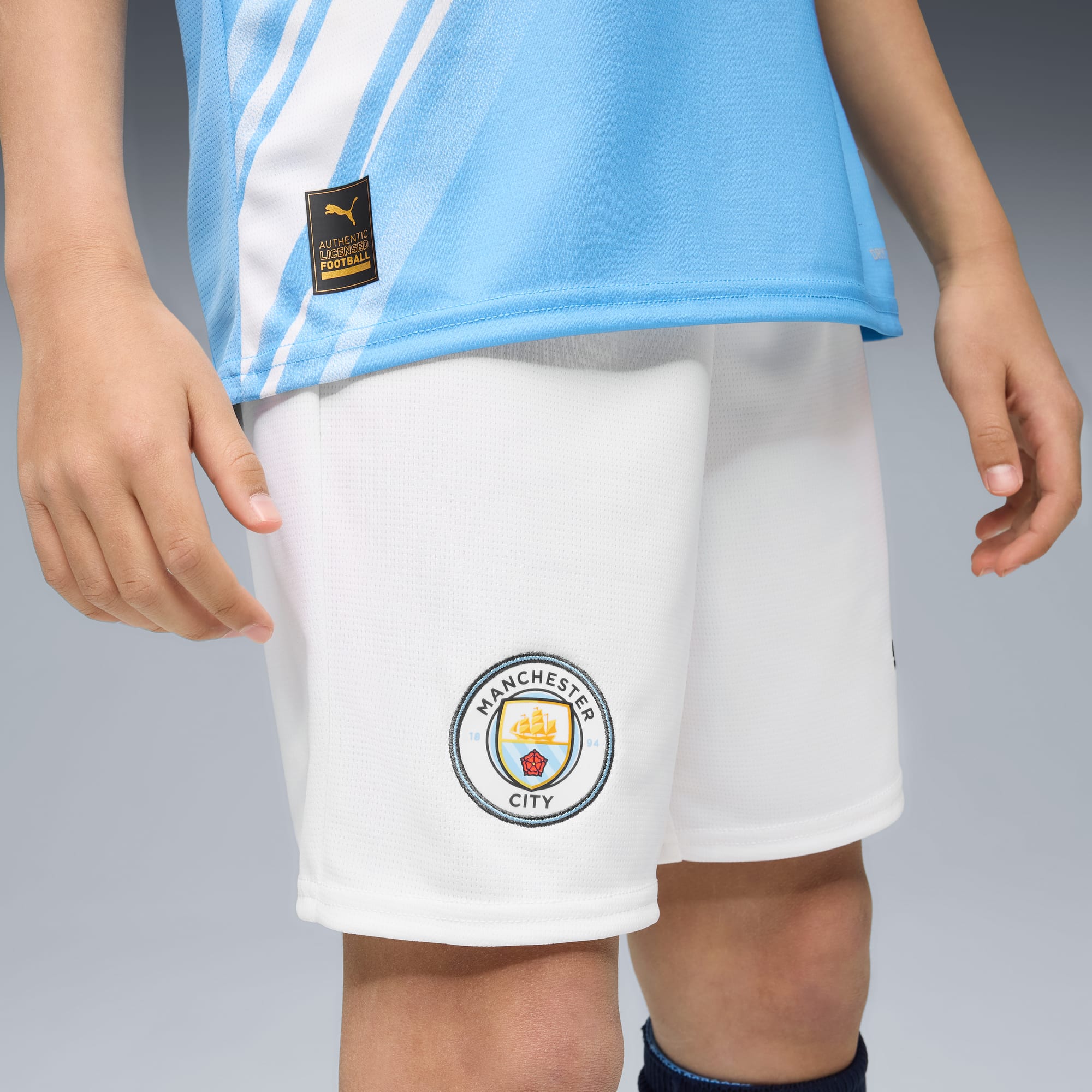 Su sfondo grigio, un modello indossa top azzurro e pantaloncini bianchi del Manchester City.