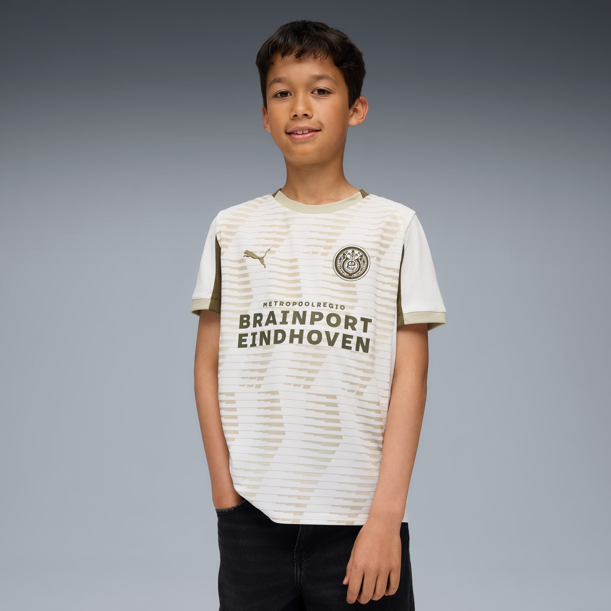 Tegen een grijze achtergrond toont een kind een wit en beige voetbalshirt en een donkere broek.