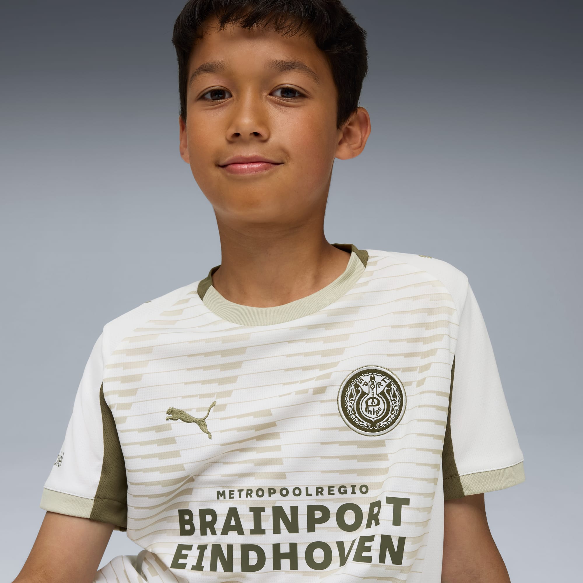 Sobre gris, niño modela camiseta de fútbol blanca y beige con logotipos.