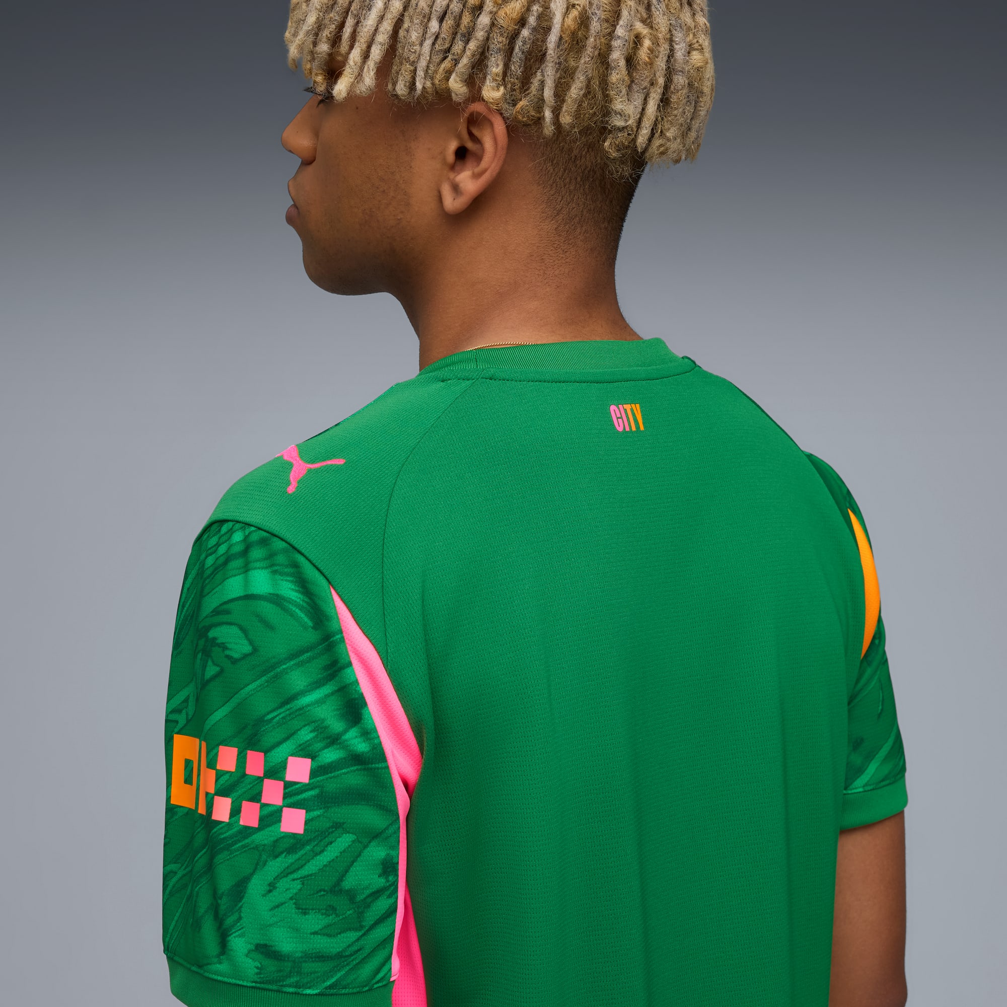 Sobre un fondo gris, un modelo viste una camiseta verde con detalles rosas y naranjas, visto desde atrás.
