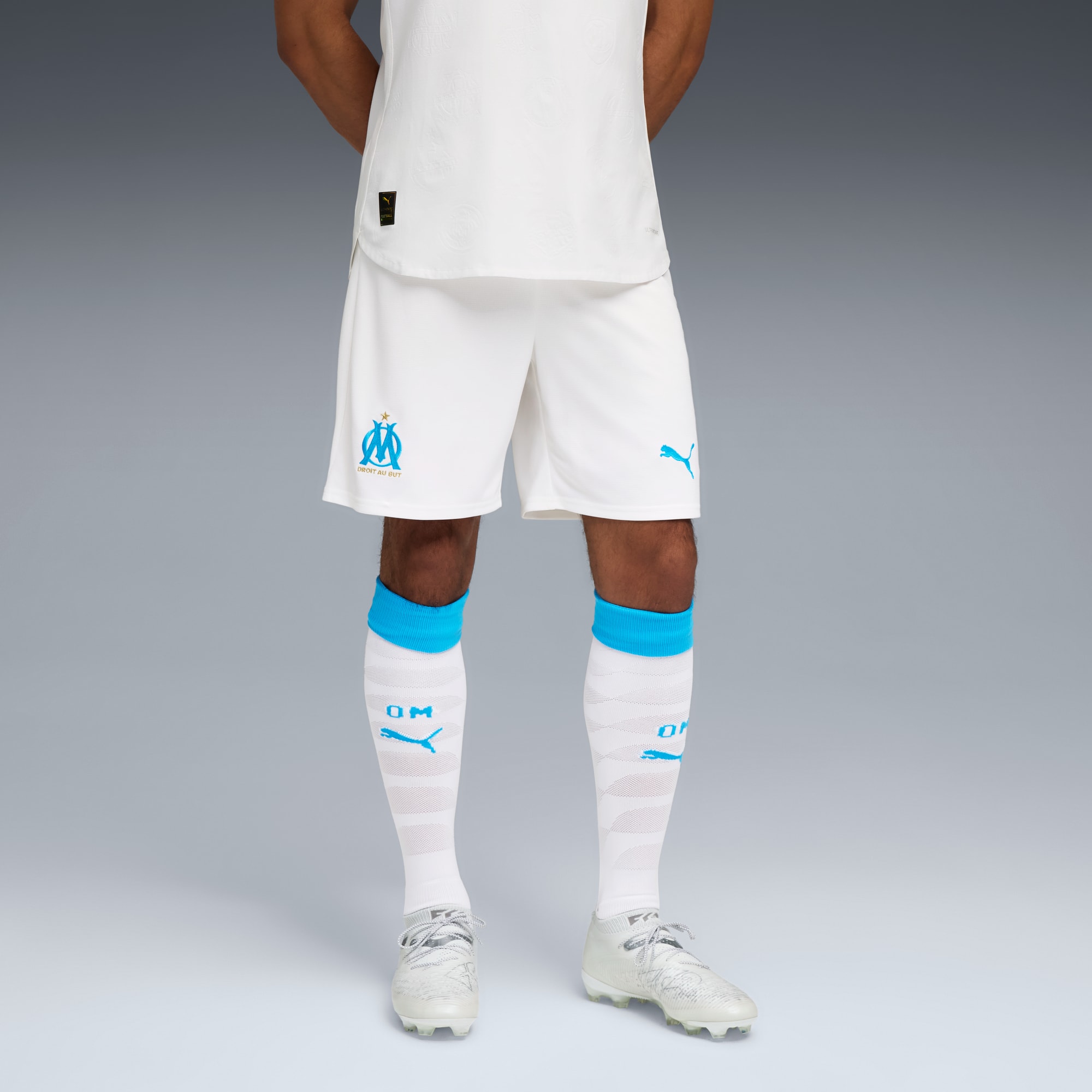 Sur fond gris, un modèle porte un t-shirt blanc, un short, des chaussettes et des chaussures avec des touches bleues.