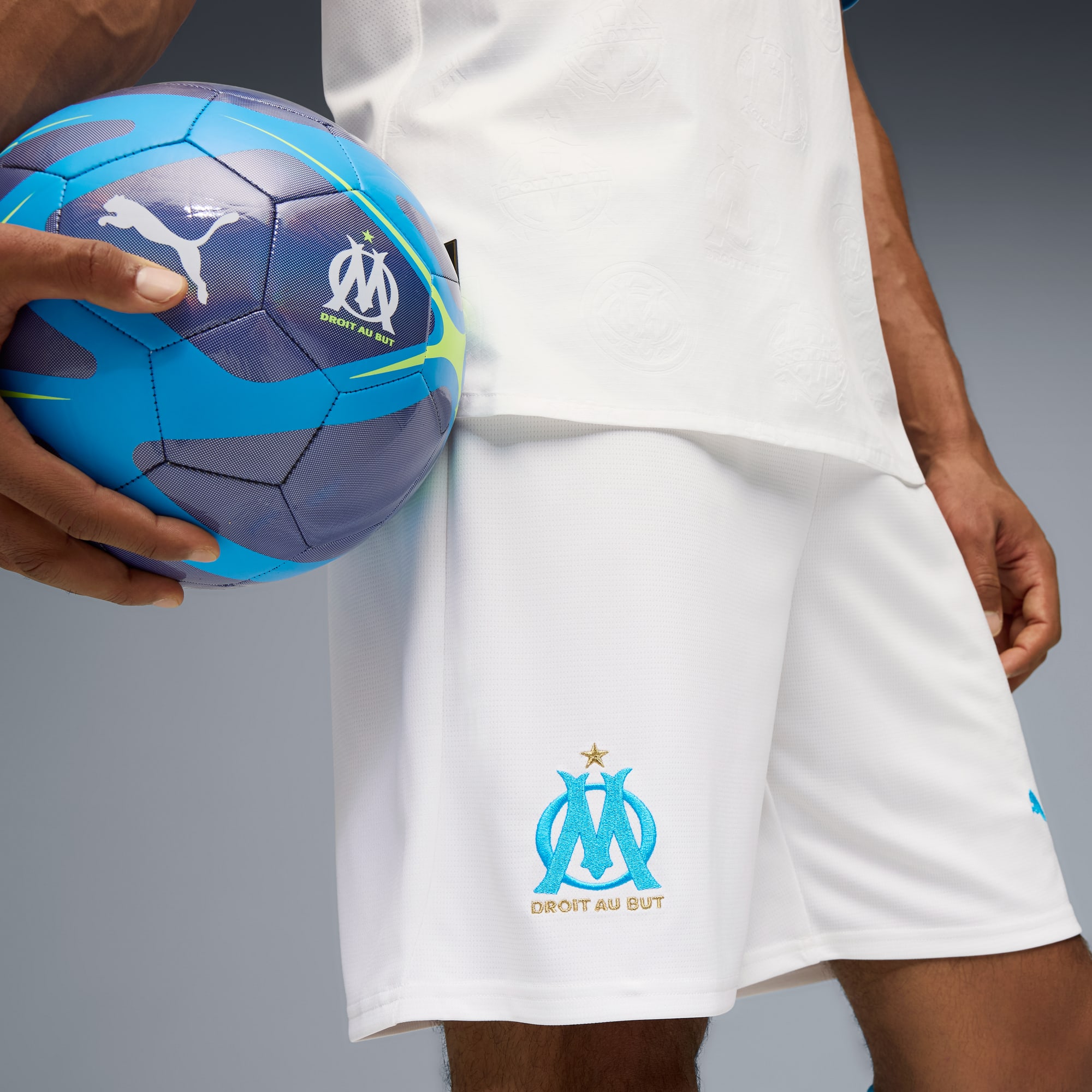 Sur fond gris, un modèle tient un ballon bleu, portant un t-shirt et un short blancs.