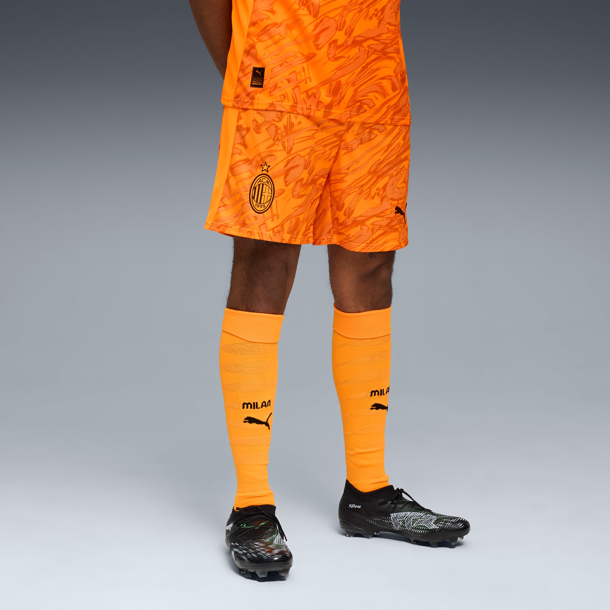 Sur fond gris, un modèle porte une tenue orange de l'AC Milan avec chaussettes et chaussures.