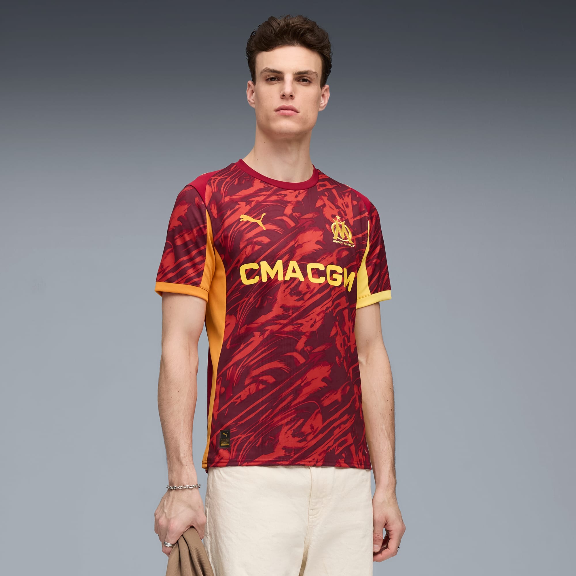 Vor grauem Hintergrund trägt ein Model ein gemustertes rotes Shirt mit gelben Akzenten und helle Hosen.