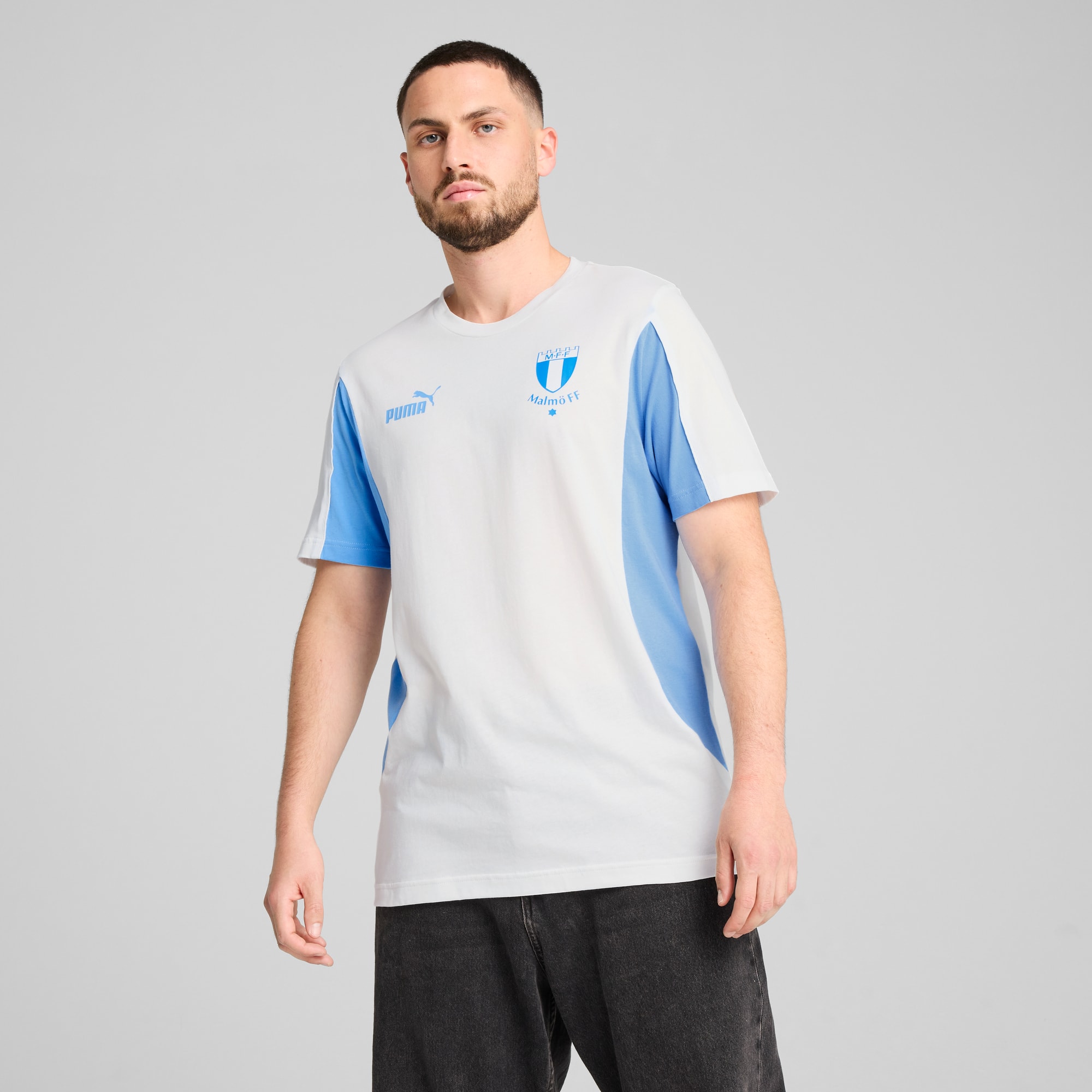 Model pozuje na szarym tle, ubrany w biało-niebieską koszulkę i ciemne spodnie.