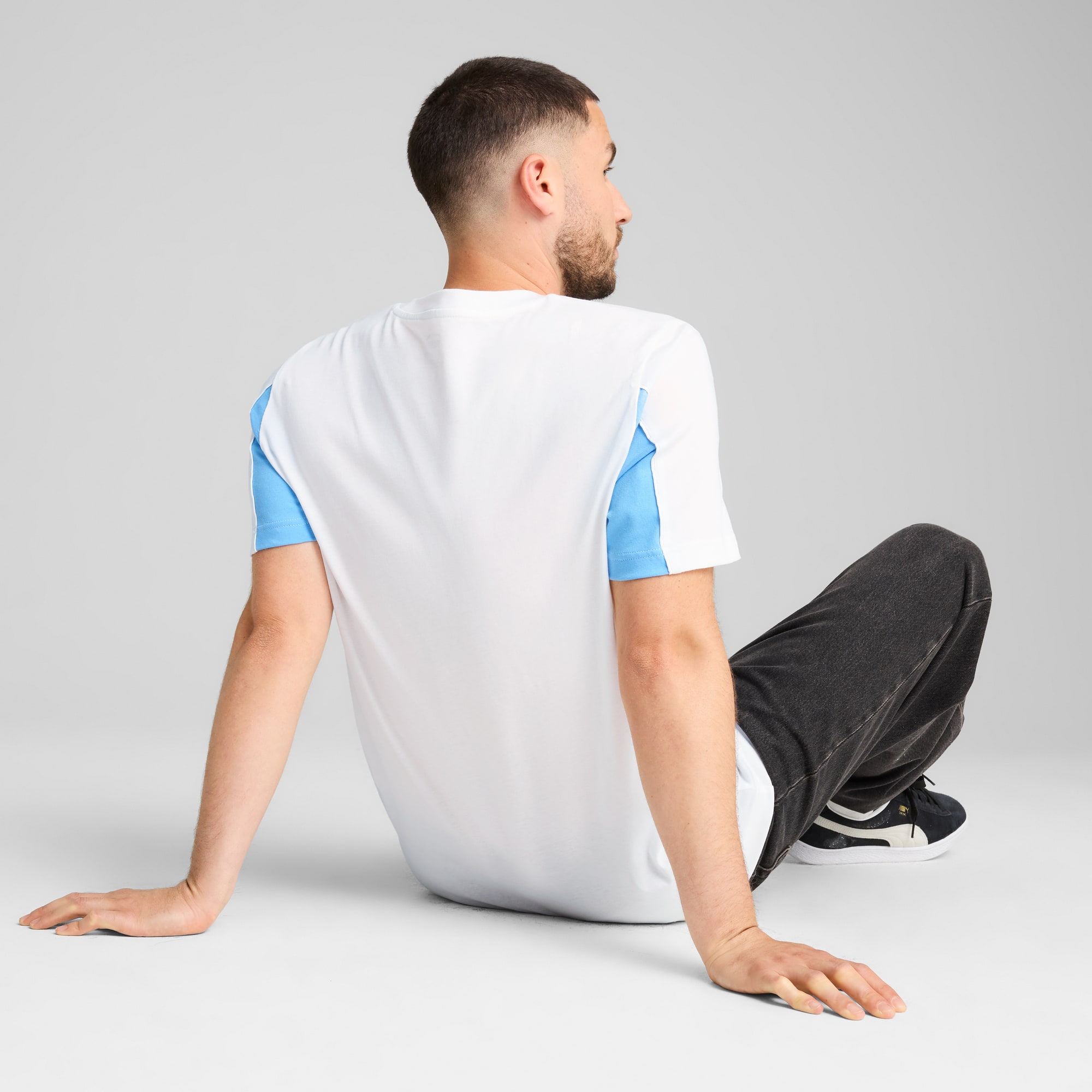 Model siedzi na szarym tle, ubrany w biało-niebieską koszulkę i ciemne spodnie.