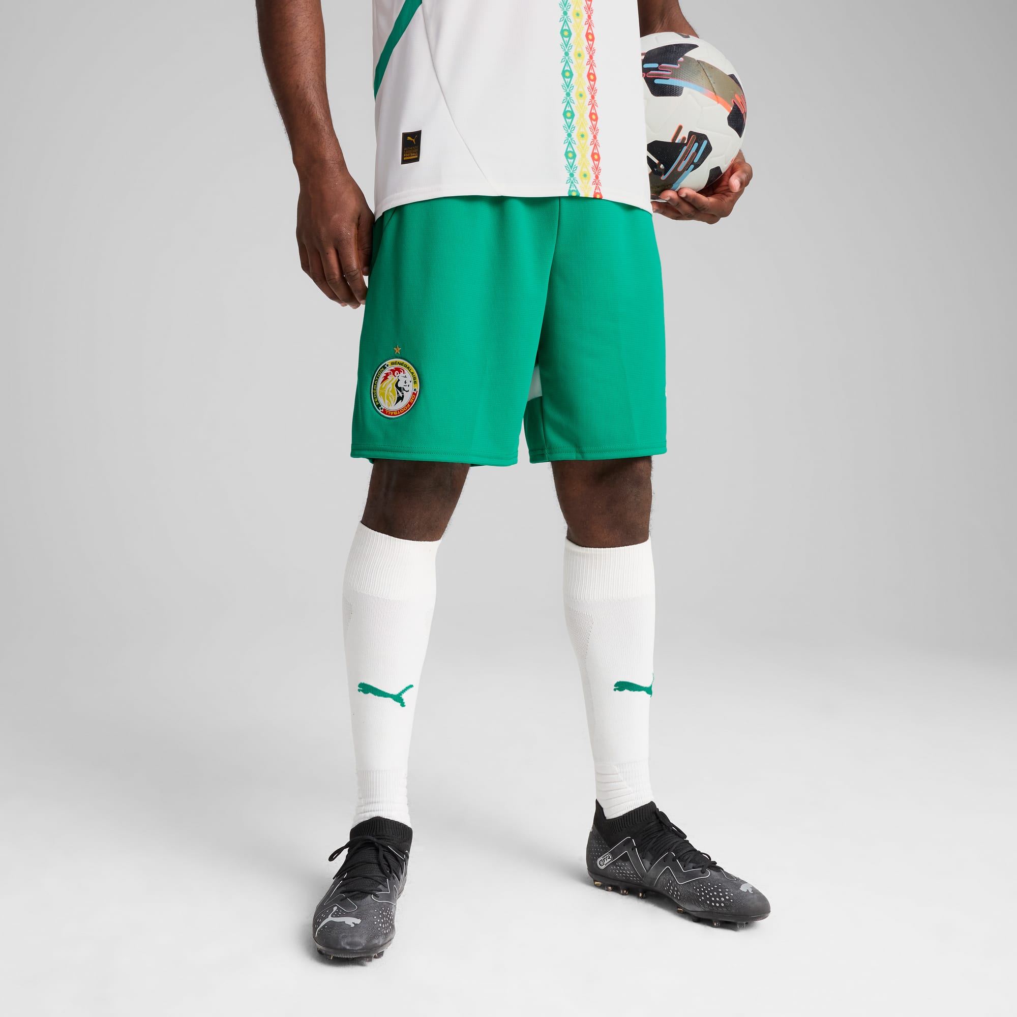 Vor grauem Hintergrund hält ein Model einen Ball, trägt ein weißes Oberteil, grüne Shorts, Socken und schwarze Fußballschuhe.