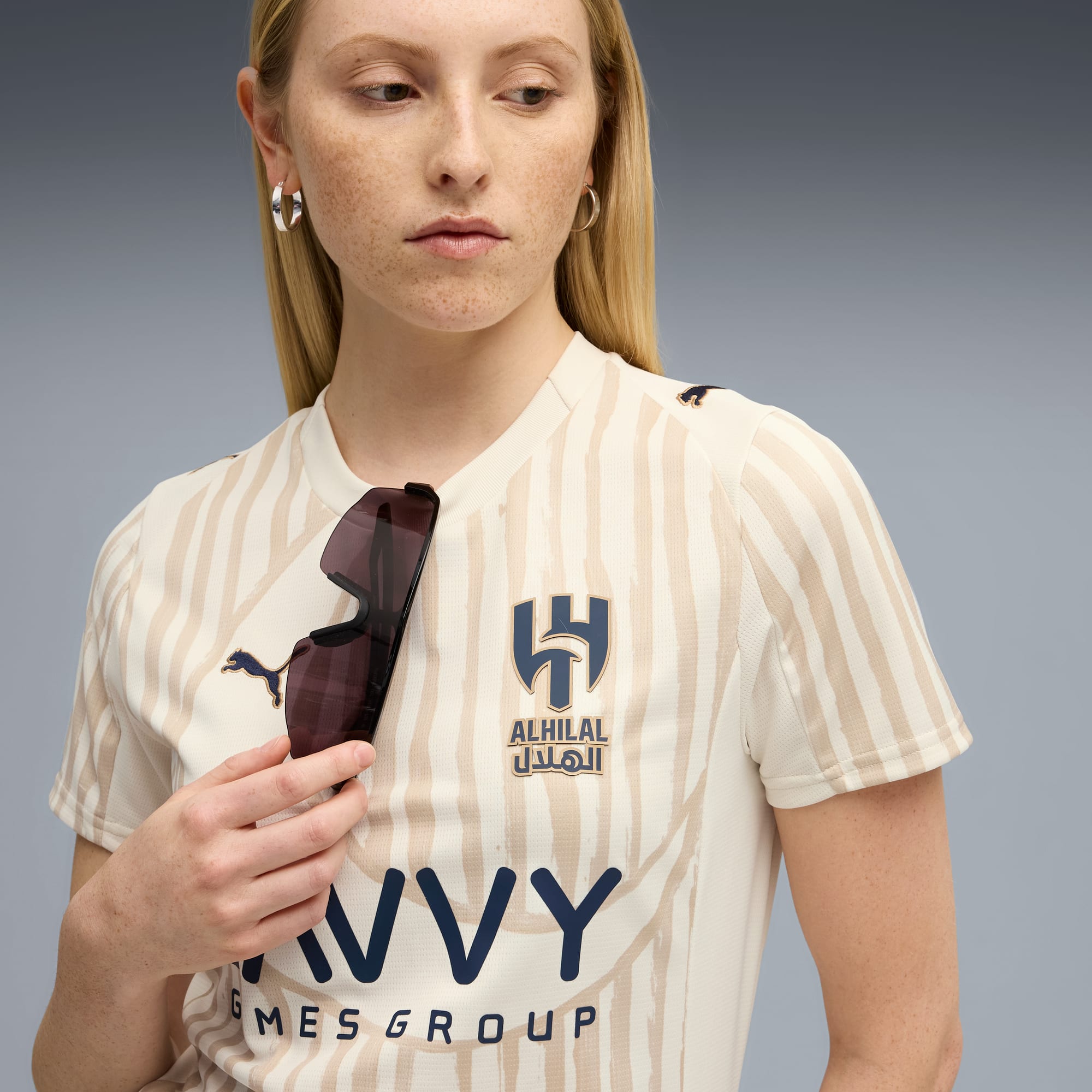 Una modella tiene degli occhiali da sole contro una maglietta a righe con un logo su sfondo grigio.