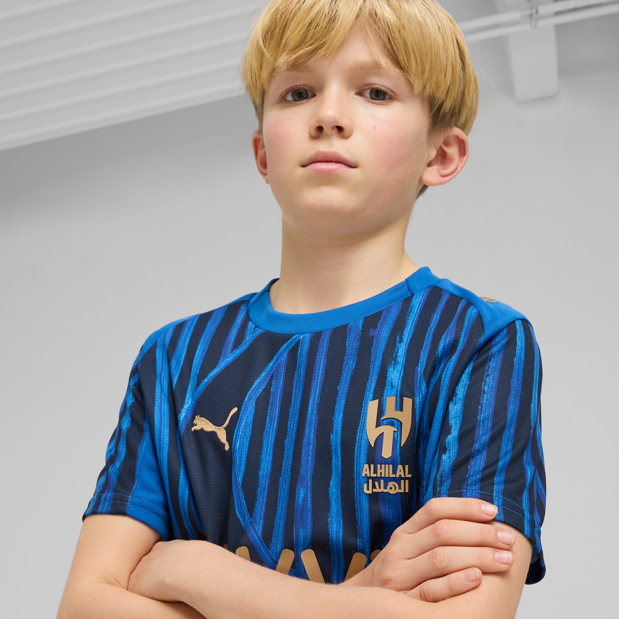 Un bambino biondo posa a braccia conserte indossando una maglia da calcio blu dell'Al Hilal.