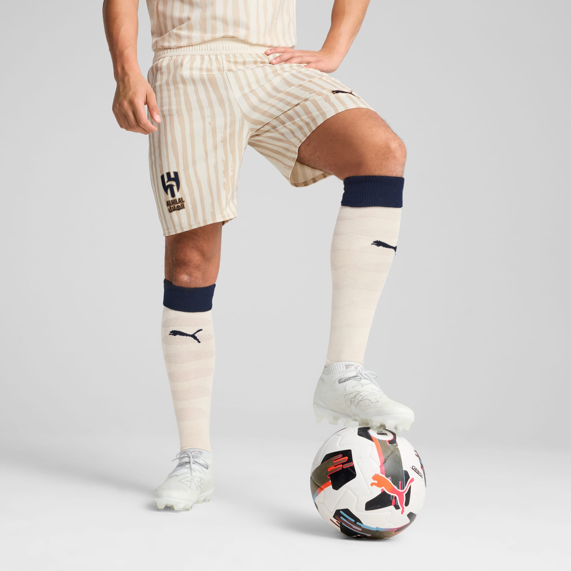Ein Model vor grauem Hintergrund, Fuß auf einem Fußball, trägt Shorts, Socken und Schuhe.