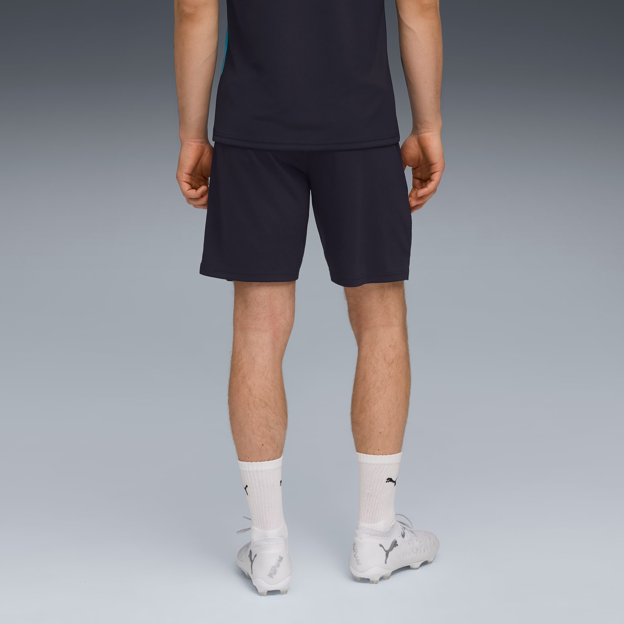 Een model staat weg van de camera op grijs, met t-shirt, donkere short, sokken en schoenen.