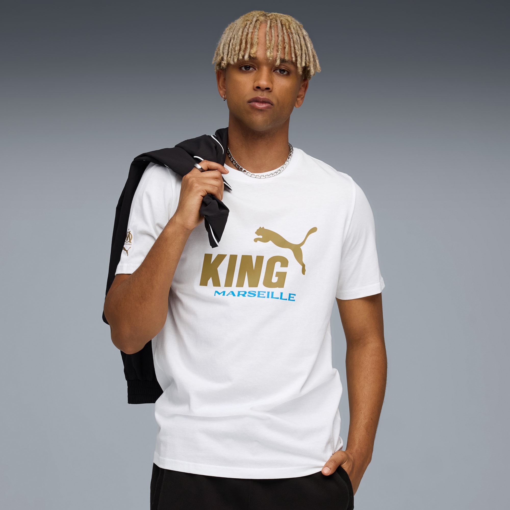 Sur fond gris, un mannequin porte un t-shirt blanc avec 'KING' en or et une veste.