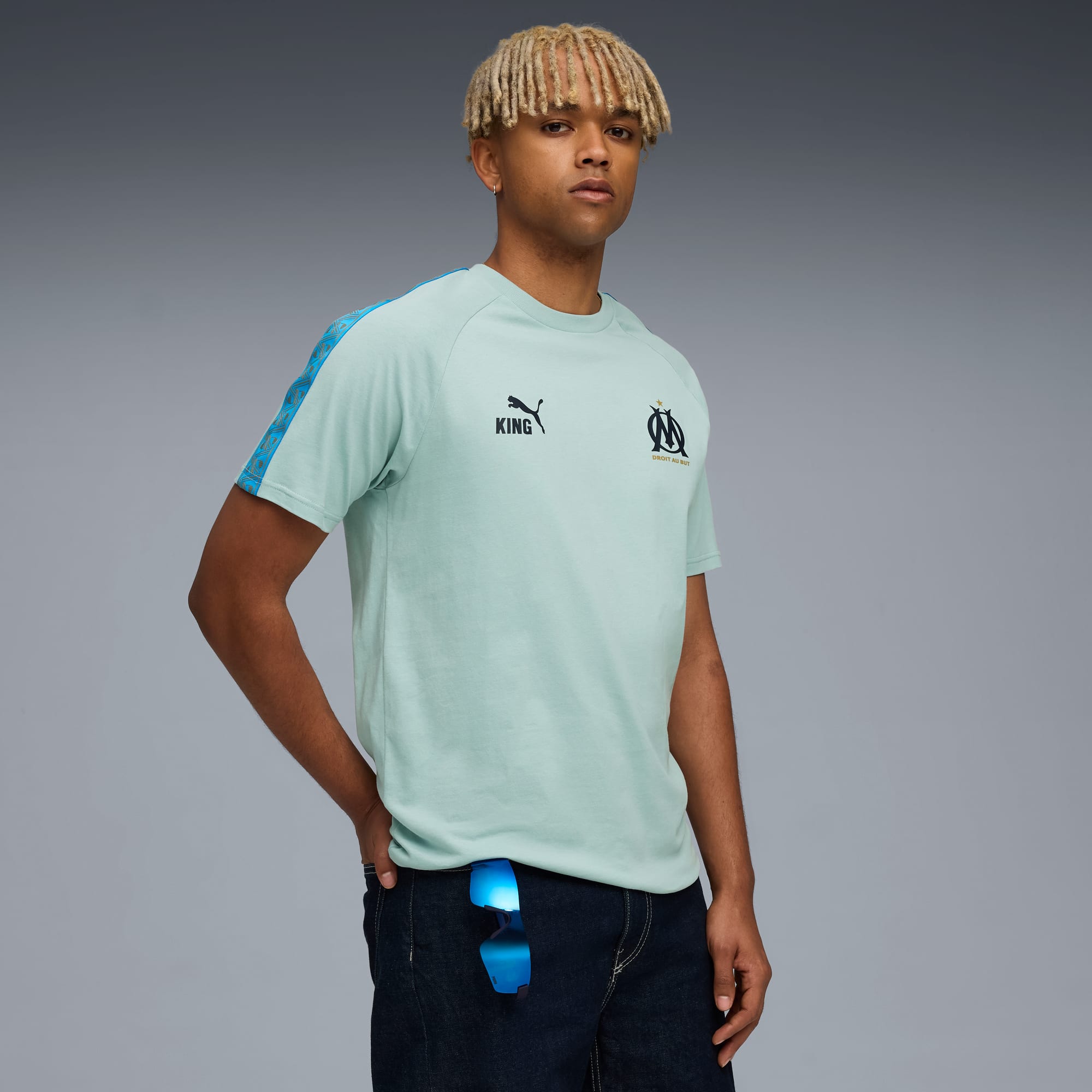Un mannequin pose sur fond gris, vêtu d'un t-shirt vert clair et d'un jean foncé, main dans la poche.
