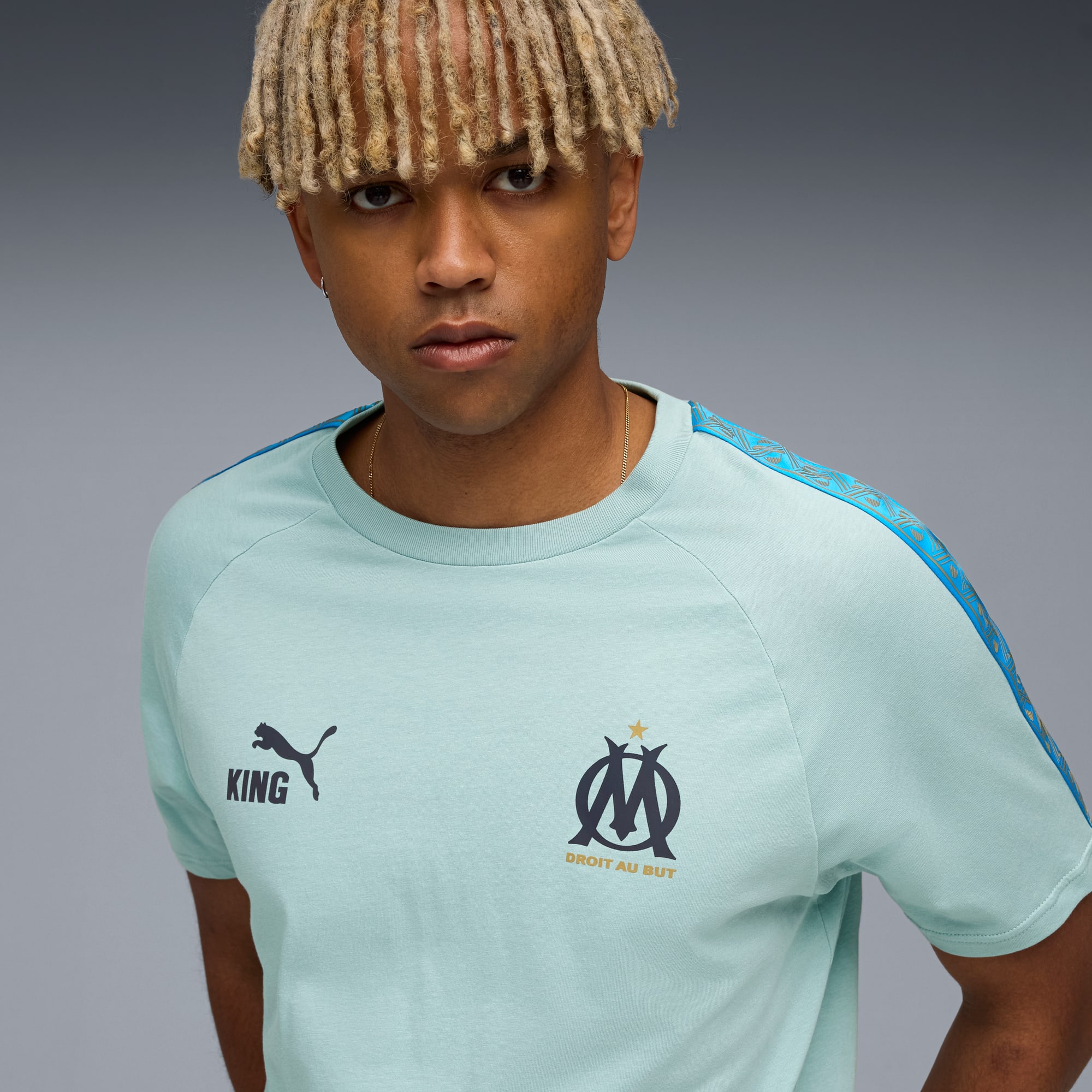 Sur fond gris, un modèle regarde devant, portant un t-shirt bleu clair avec logos.