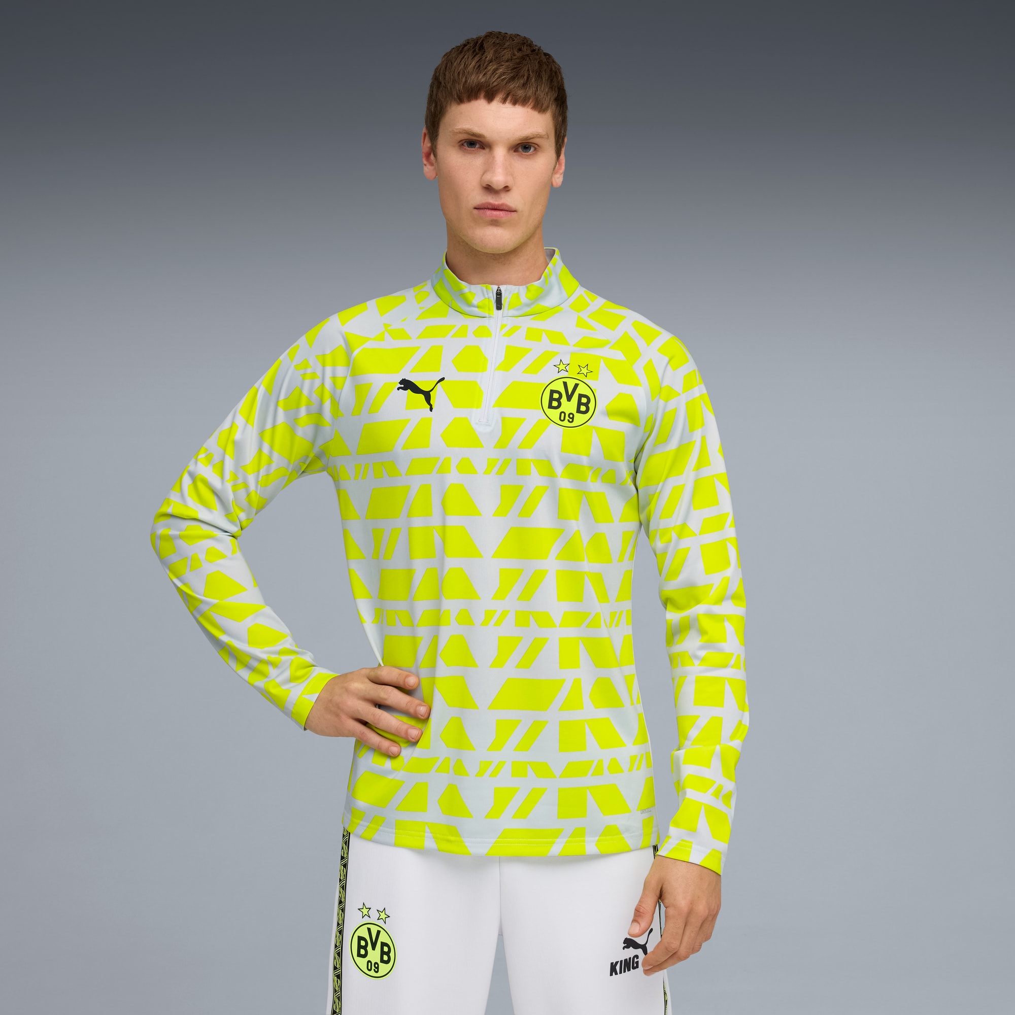 Een model poseert tegen grijs, gekleed in een geel patroontop en witte broek.