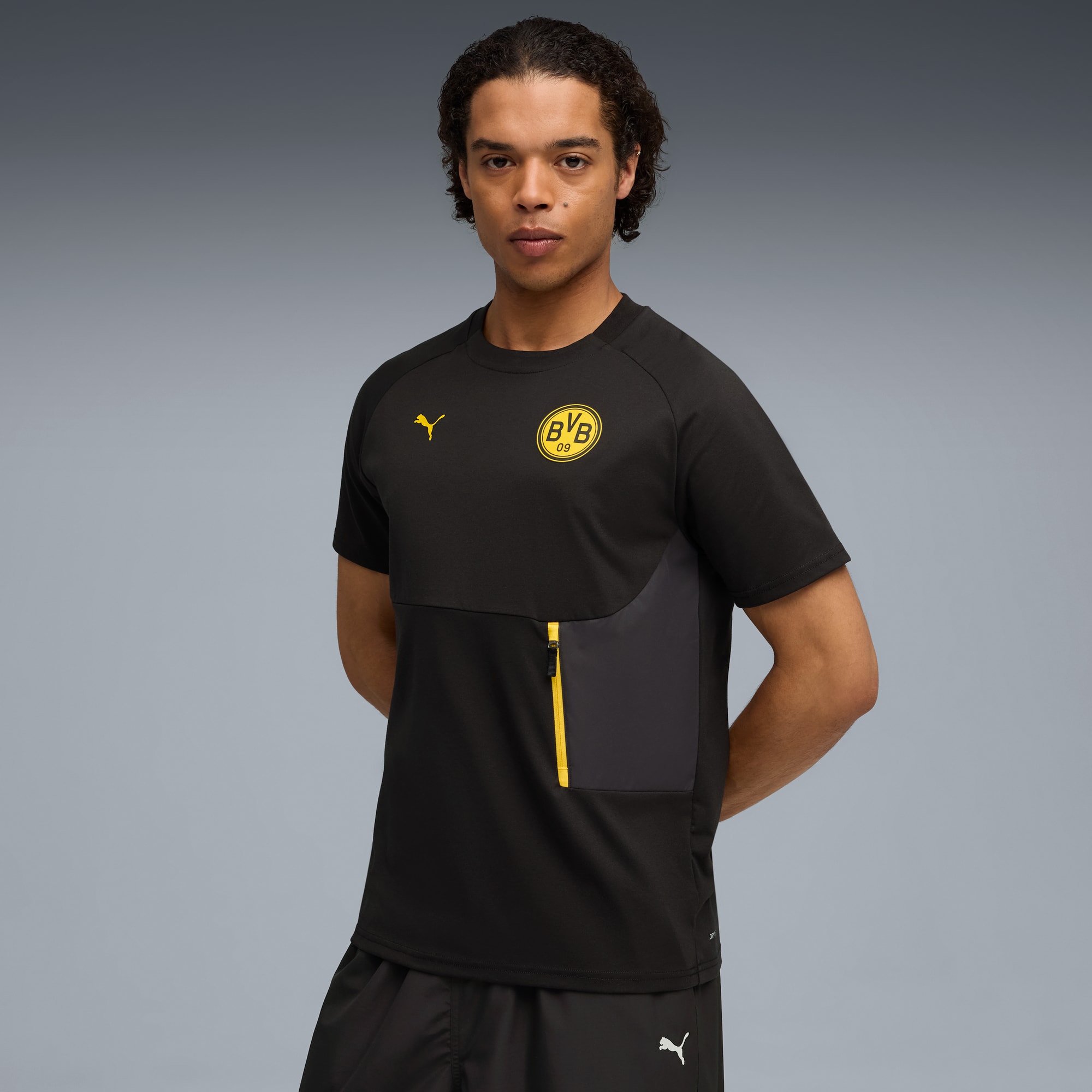 Tegen grijs draagt een model een zwart BVB-shirt en zwarte korte broek, poserend.