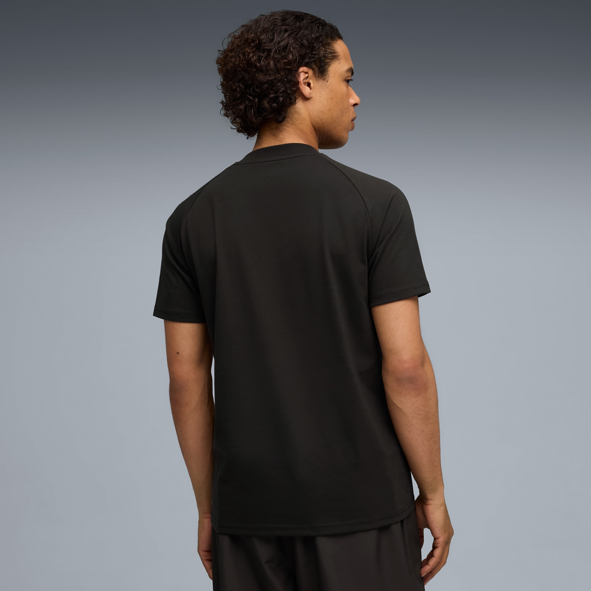 Een model staat met de rug naar de camera, draagt een zwart t-shirt en donkere short.