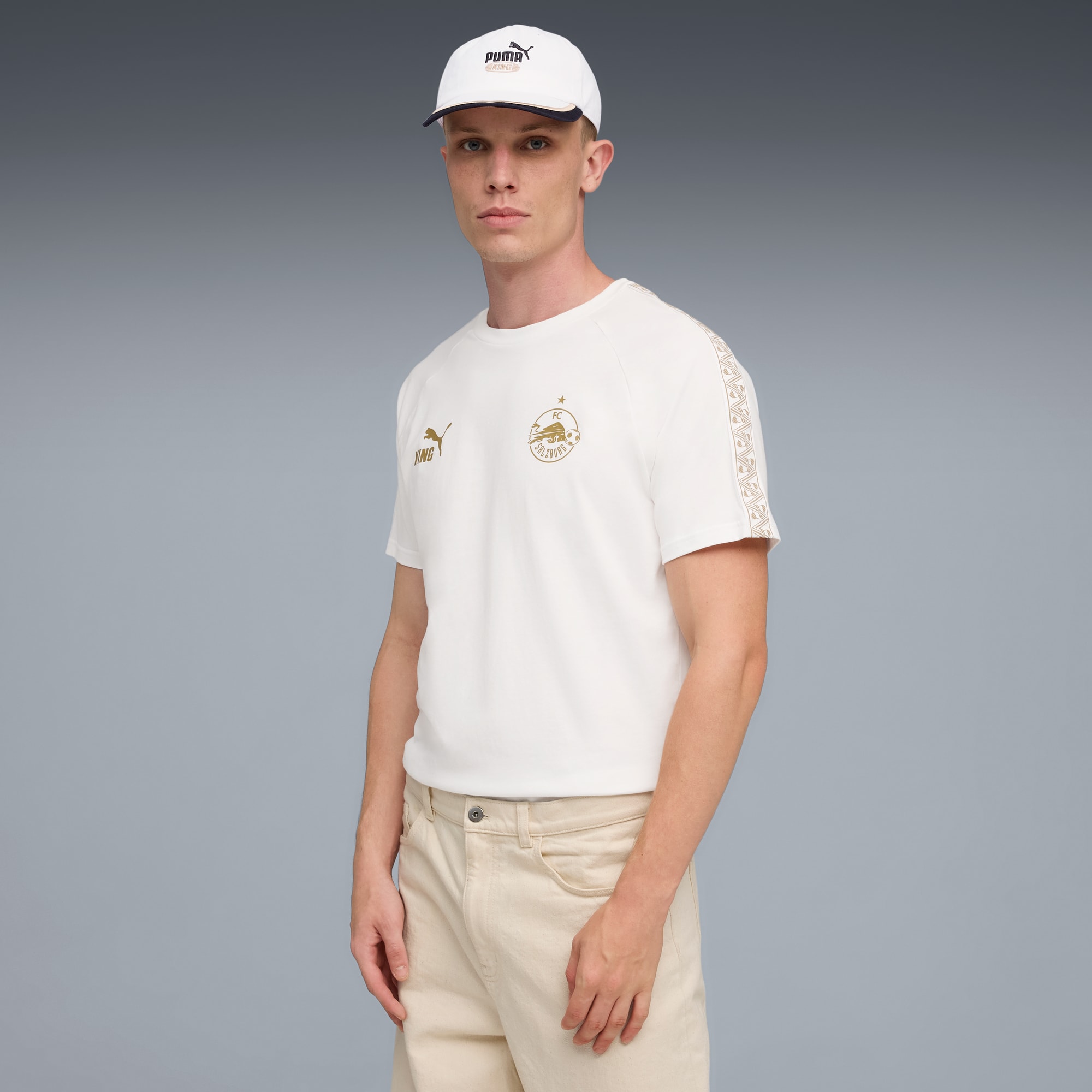 Een model staat tegen een grijze achtergrond met een wit t-shirt, pet en lichte broek.