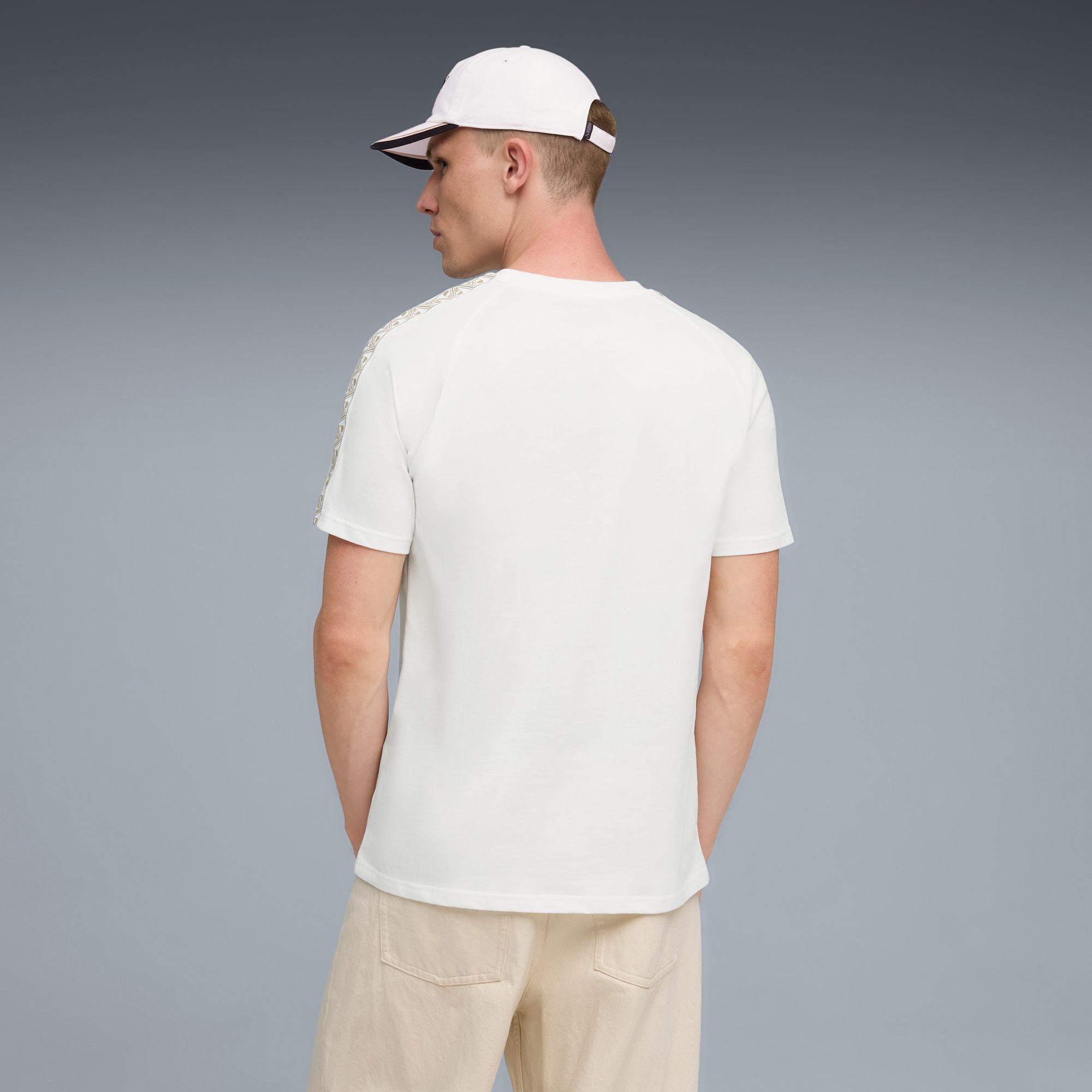 Een model staat tegen een grijze achtergrond, gekleed in een wit T-shirt, beige shorts en een witte pet.