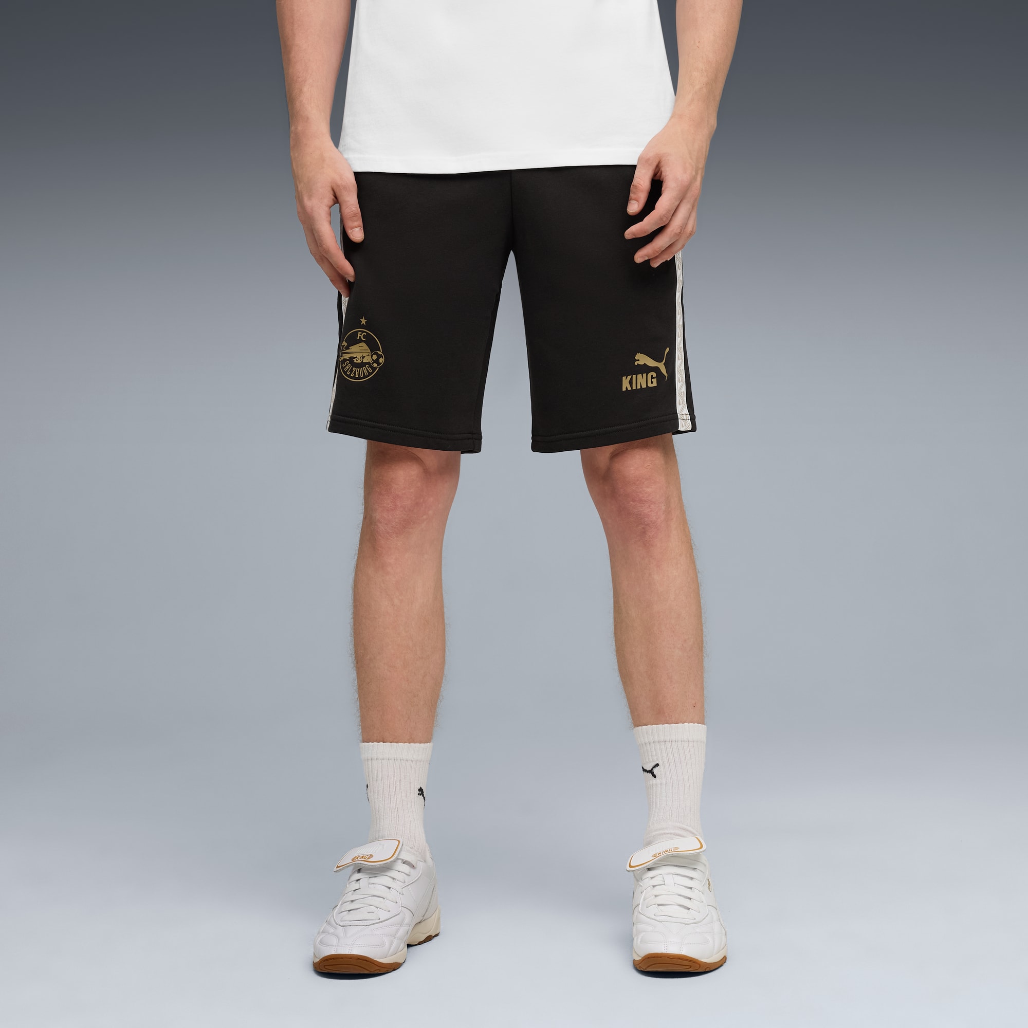 Un mannequin sur fond gris porte un t-shirt blanc, un short noir avec logos dorés et des baskets blanches.