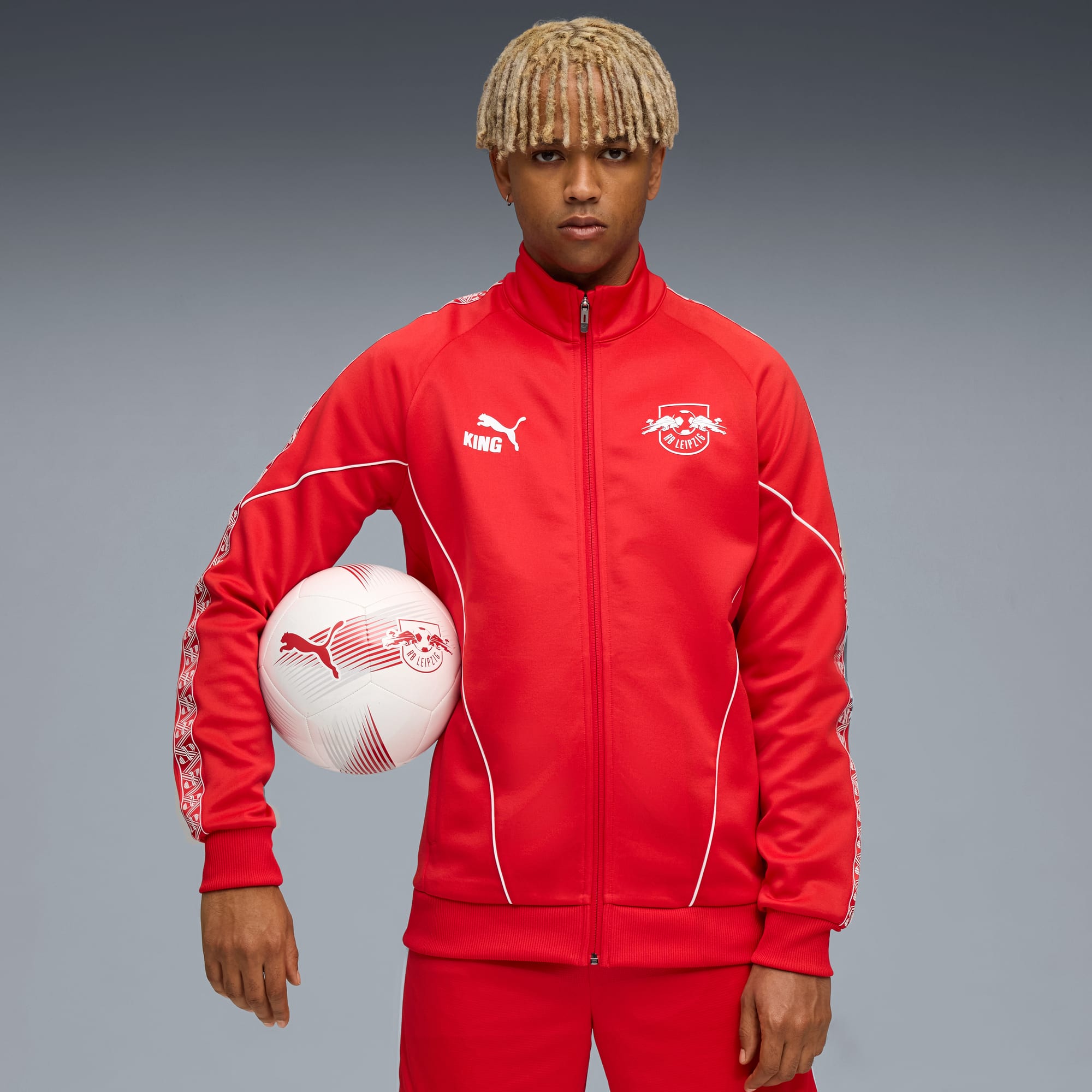 Tegen grijs houdt een model een voetbal vast, gekleed in een rood trainingspak.
