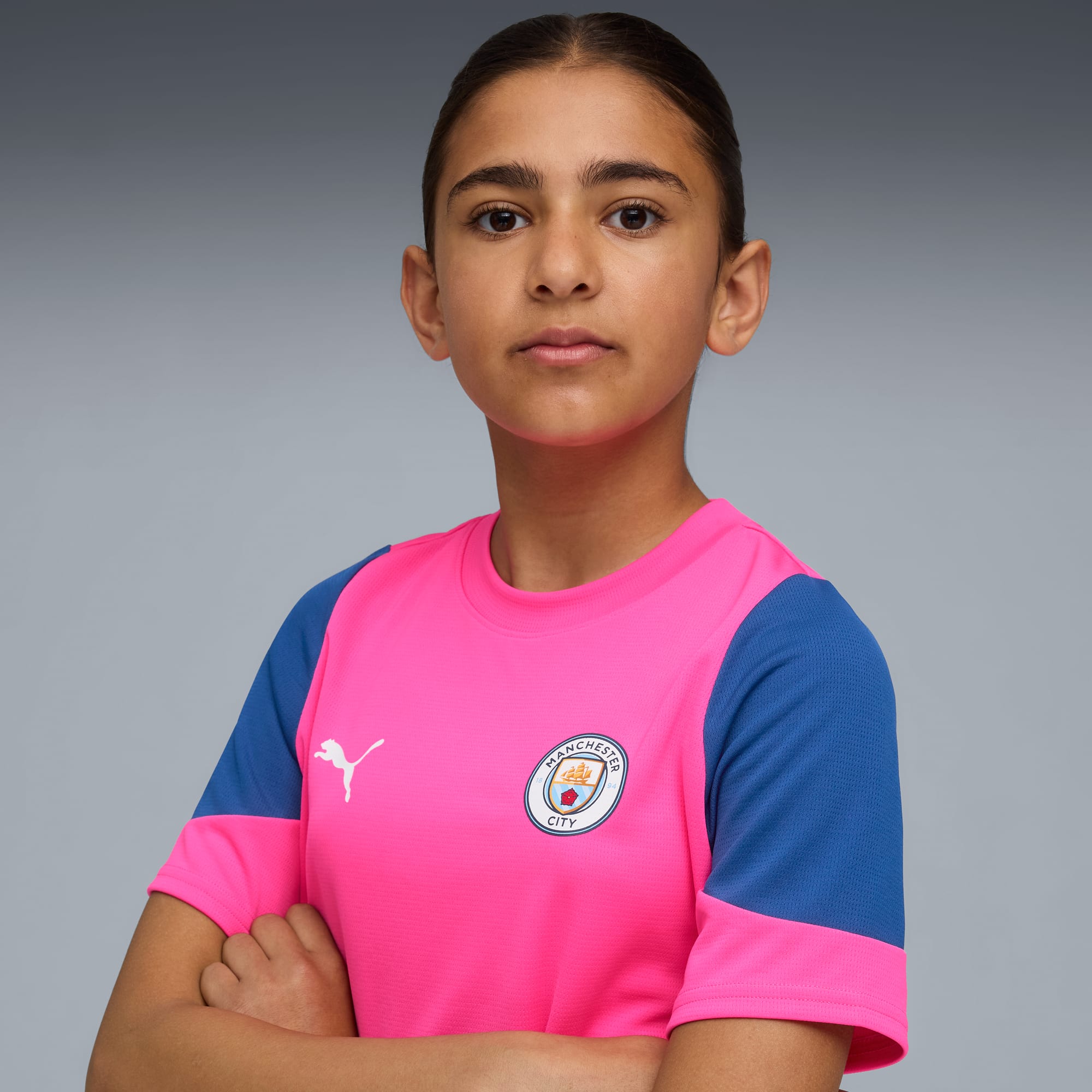 Against grey, a model stands with arms crossed, wearing a pink and blue Manchester City t-shirt.