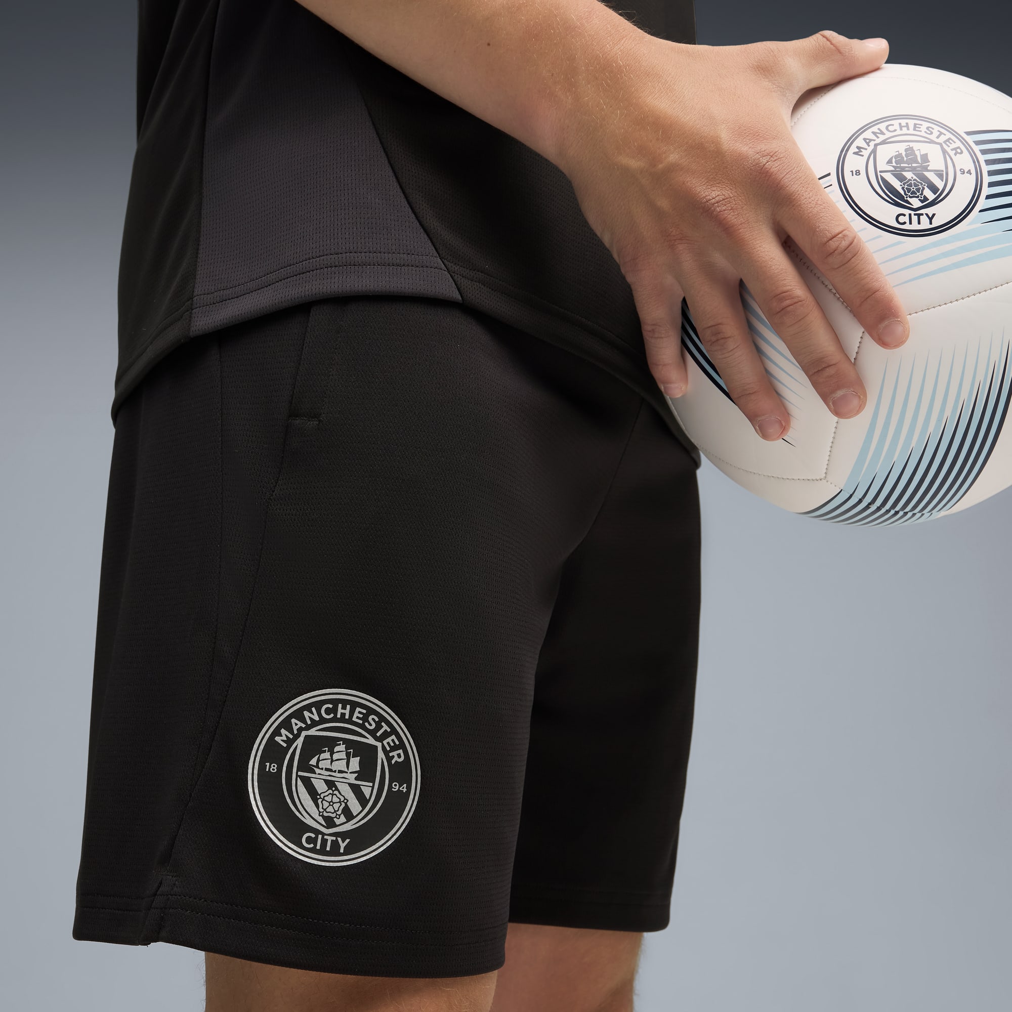 Su sfondo grigio, una persona tiene in mano un pallone da calcio del Manchester City e indossa pantaloncini neri.