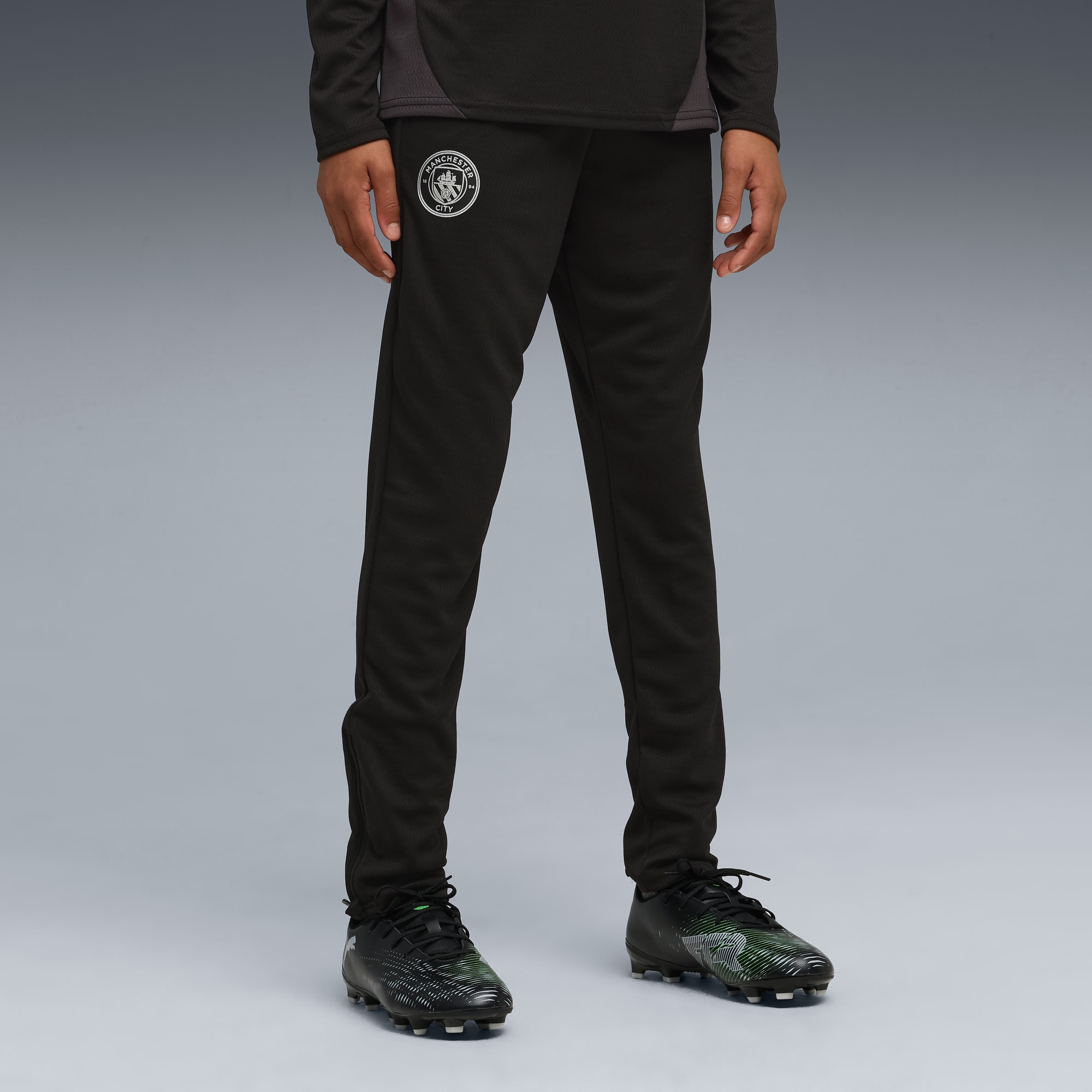 Sur fond gris, un modèle porte un pantalon de survêtement noir et des chaussures de foot.