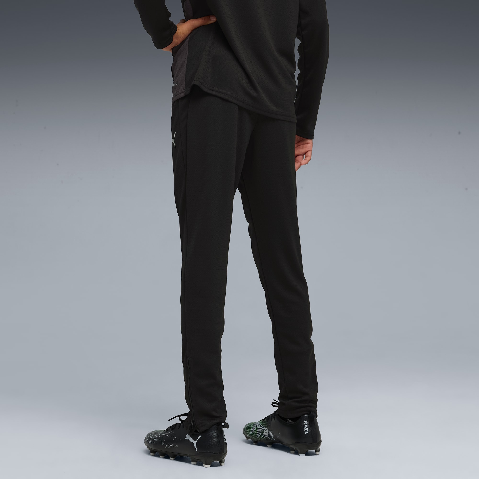 Un modèle sur fond gris porte un pantalon noir et des chaussures de foot, vu de dos.