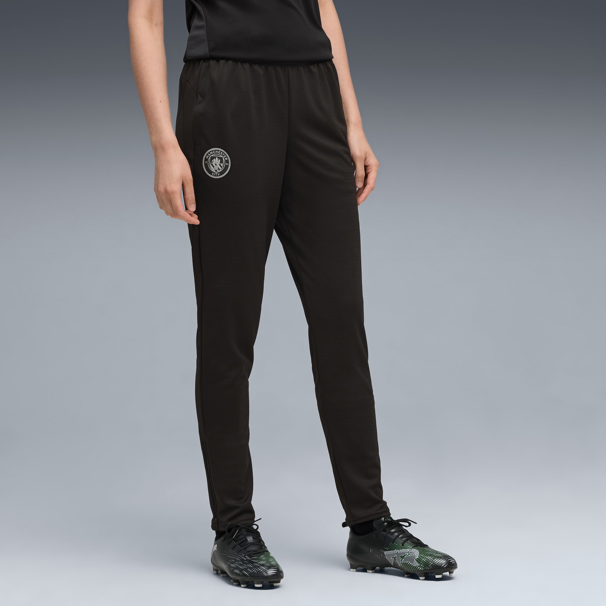 Su sfondo grigio, un modello indossa pantaloni da jogging neri con logo e scarpe da calcio abbinate.
