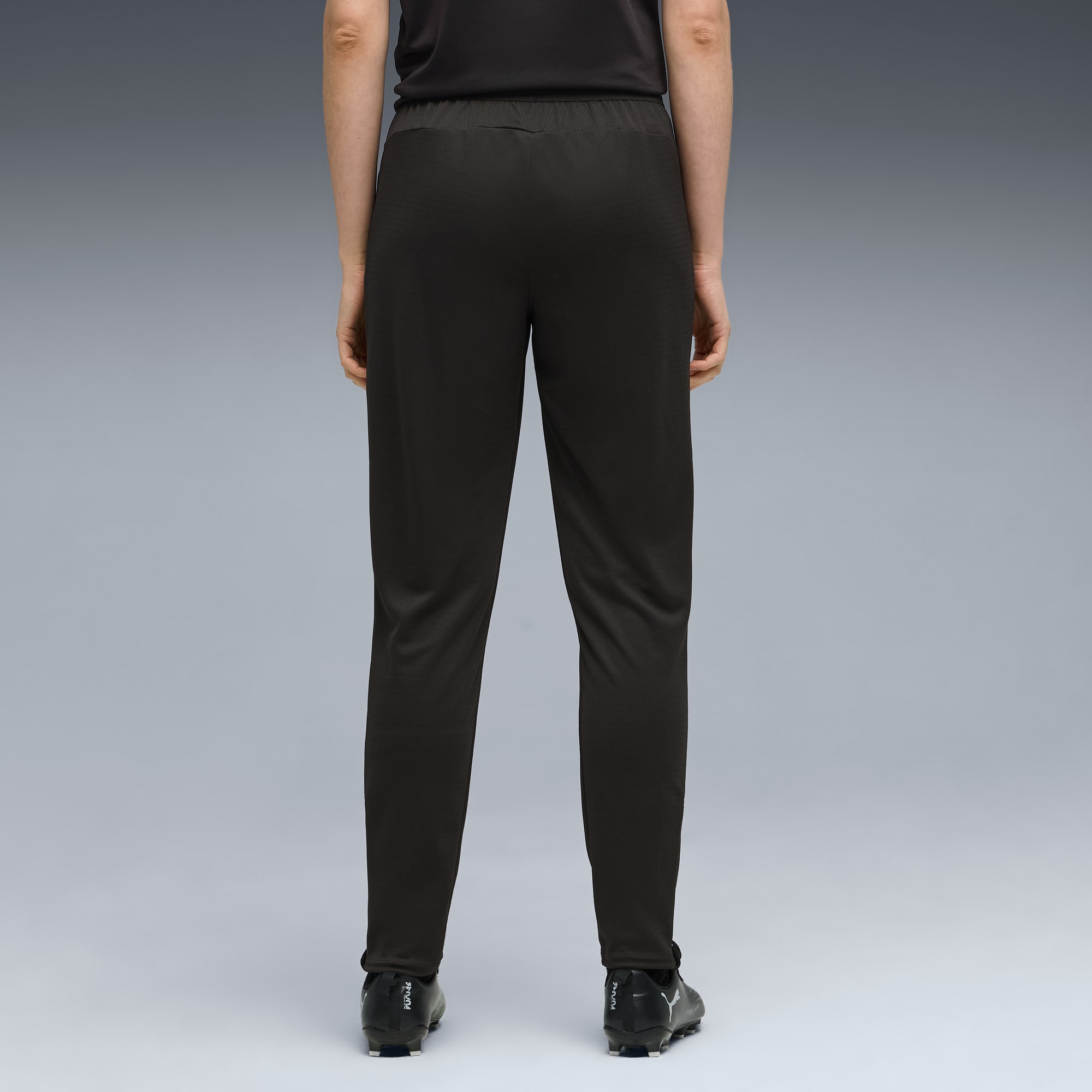 Een model staat op een grijze achtergrond, van achteren gezien, in zwarte broek en voetbalschoenen.